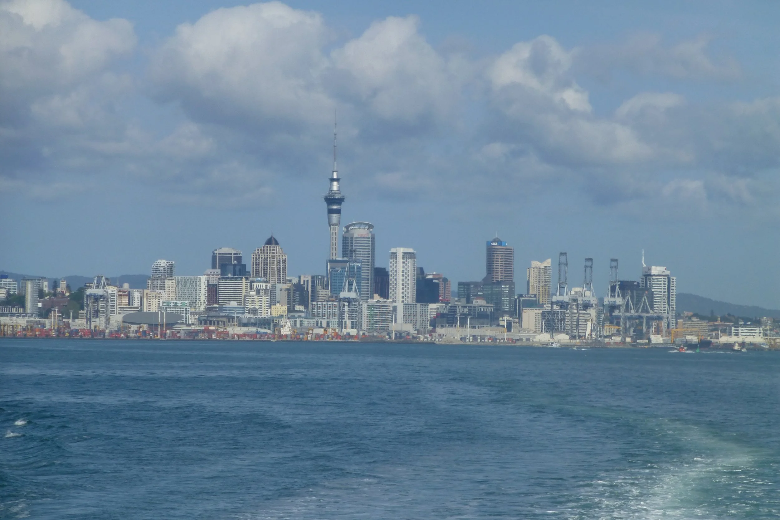 View of the Auckland city skyline with tall buildings and the prominent sky tower, seen from across a body of water.