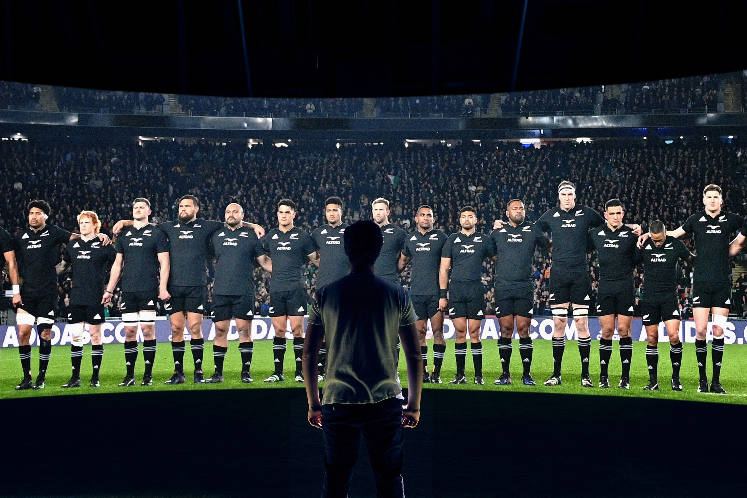 A person standing with their back to the camera facing the national rugby team the All Blacks performing a maori war dance the haka.