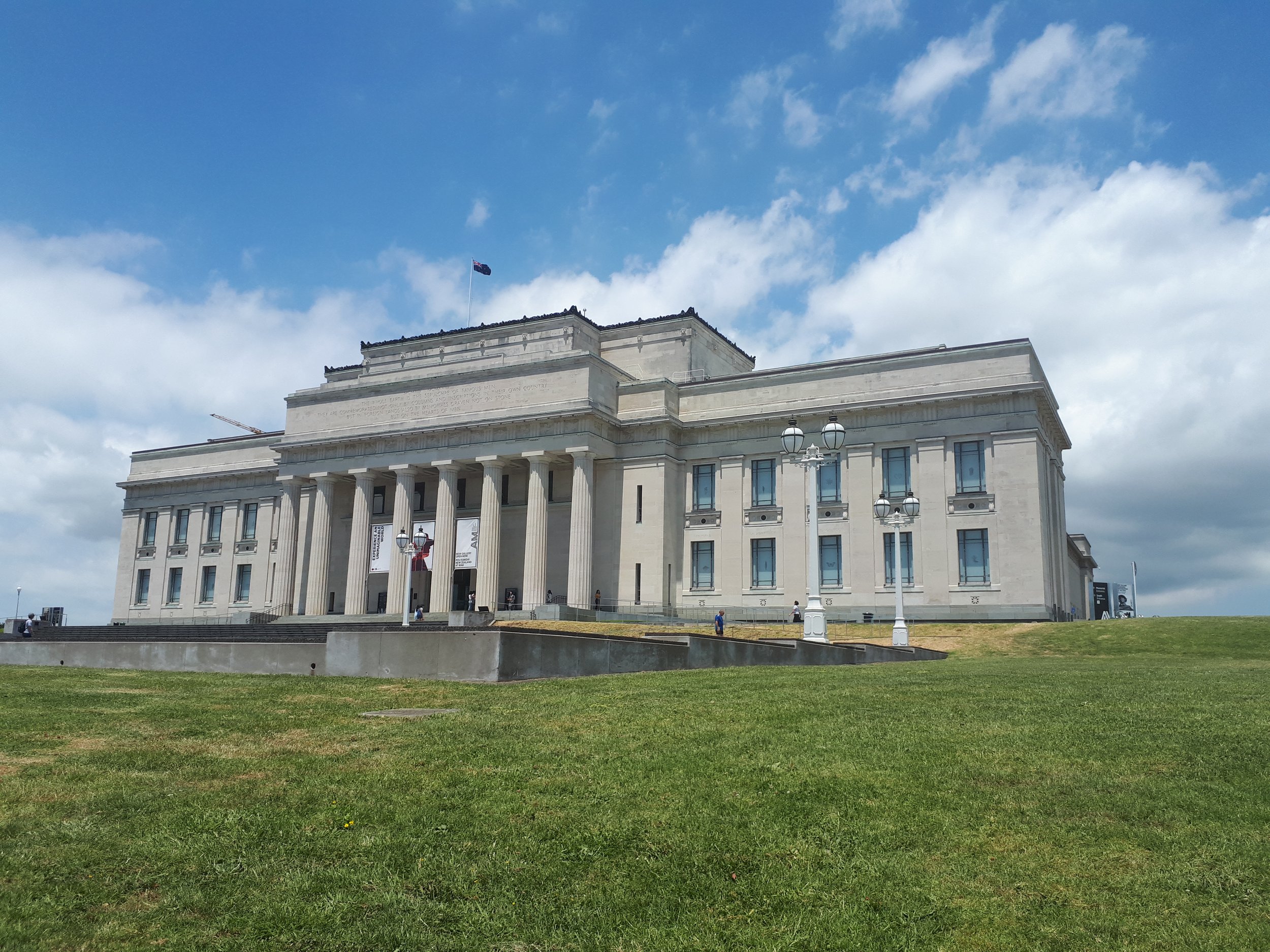 A classical-style building, the Auckland War Memorial Museum, with large columns and steps, surrounded by a grassy area of the Auckland Domain and street lamps, under a partly cloudy sky.