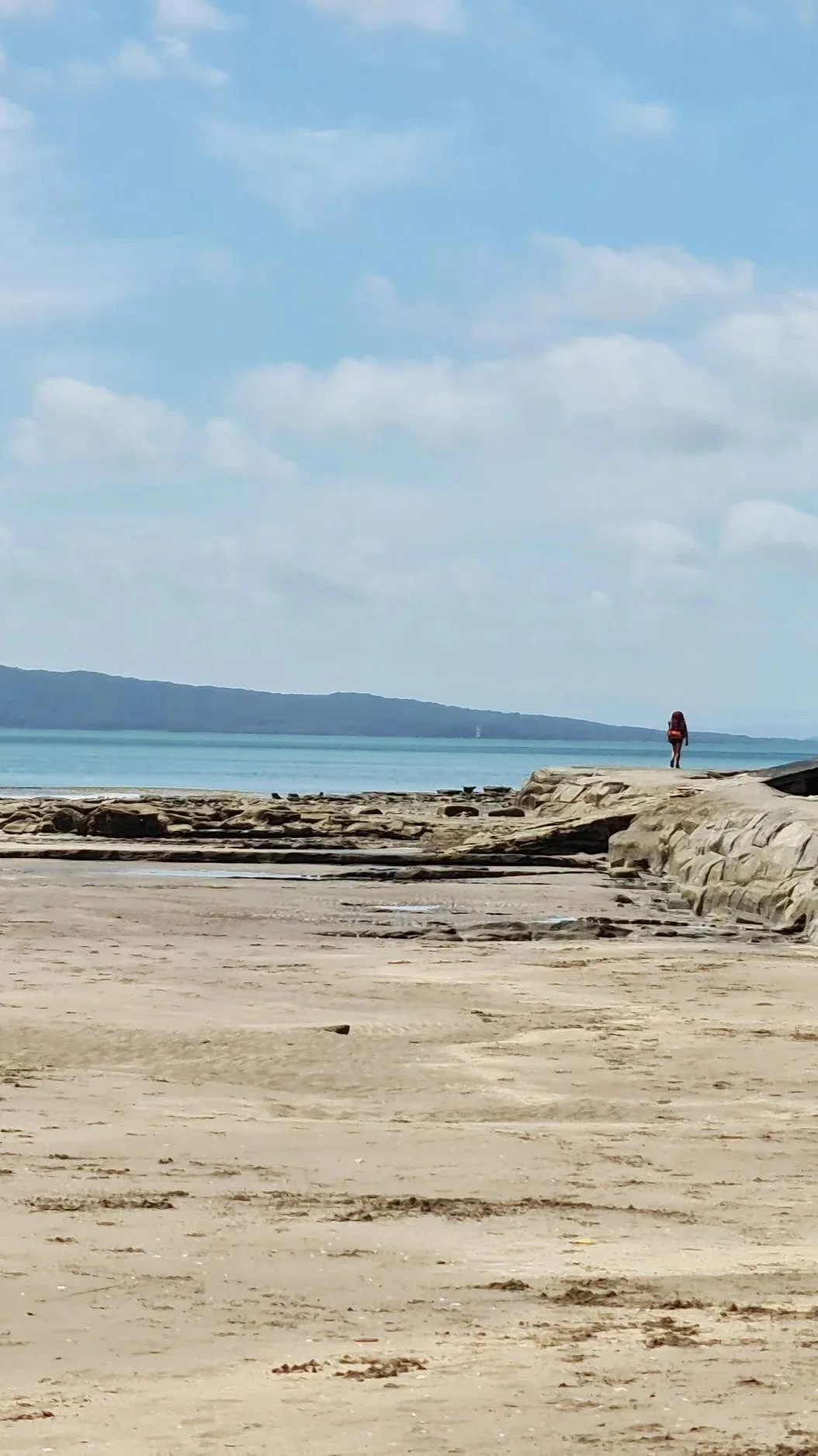 Walking a coastal Auckland section of the Te Araroa Trail with beach, sand and rocks, a person is walking near the shoreline, calm blue water, and a partly cloudy sky.