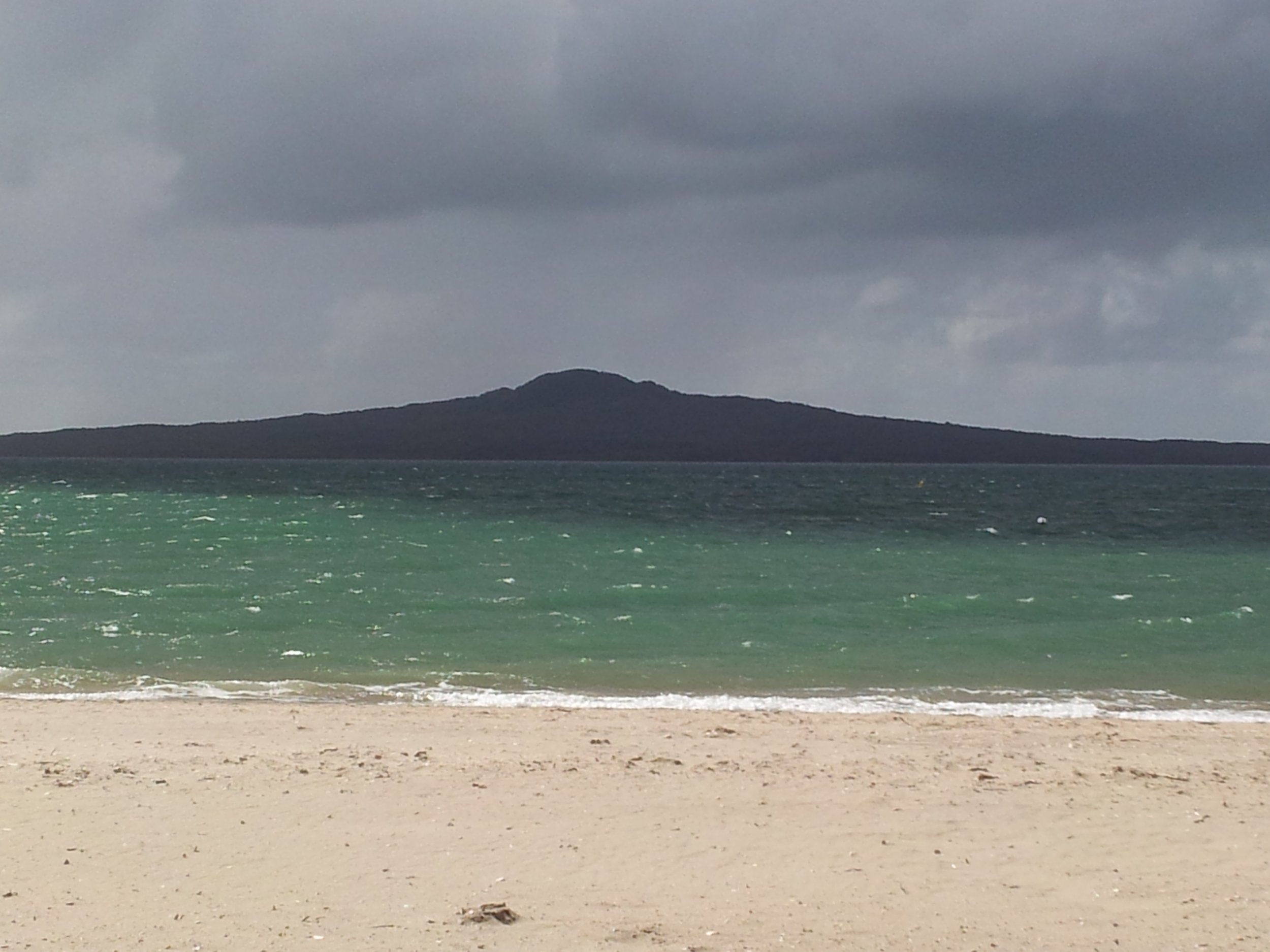 A beach with light-colored sand, greenish ocean water, and a mountainous island Rangitoto in the distance under a cloudy sky.