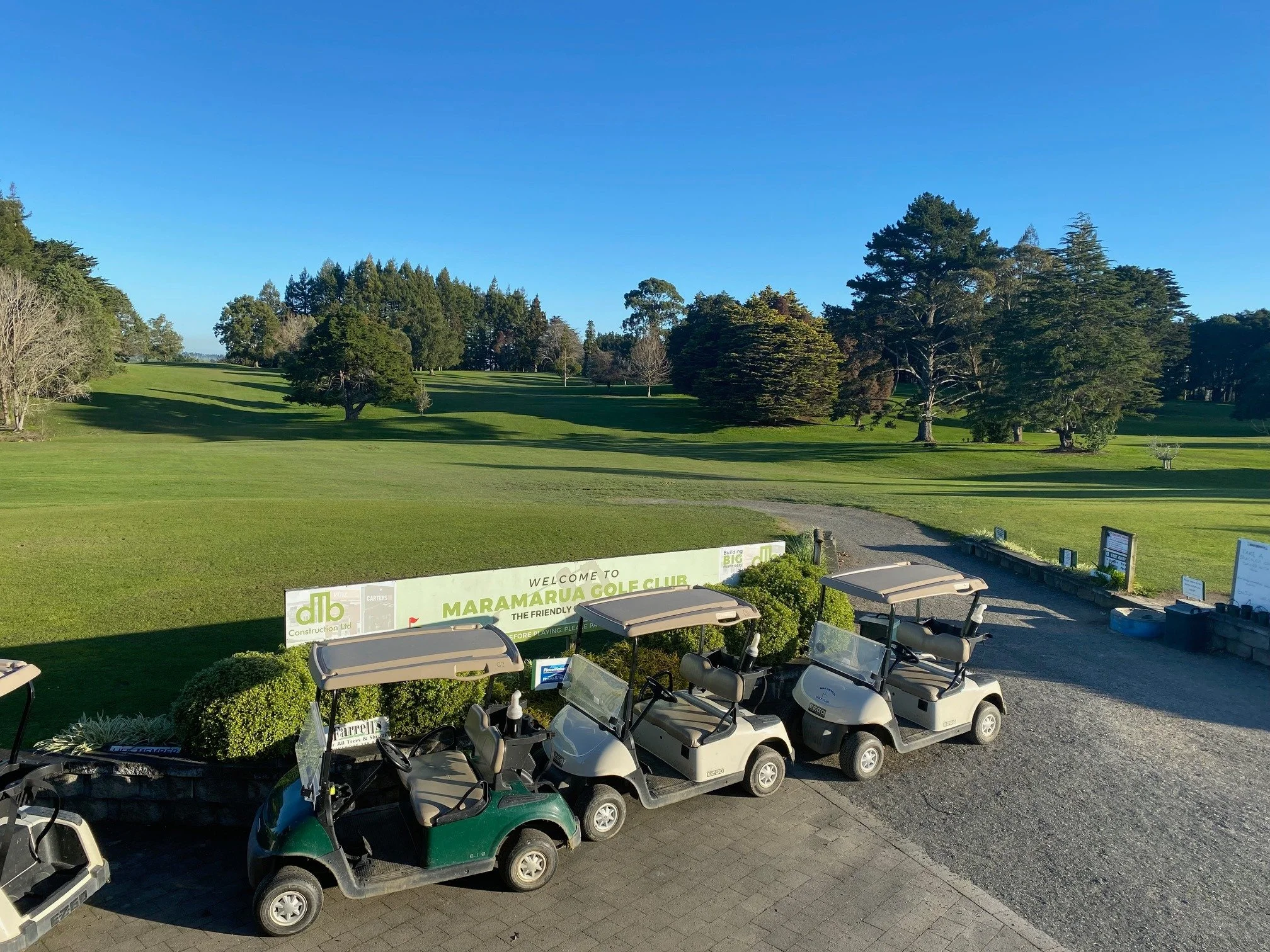 Golf carts parked near a sign at Maramarua Golf Club, with green trees and a clear blue sky in the background.
