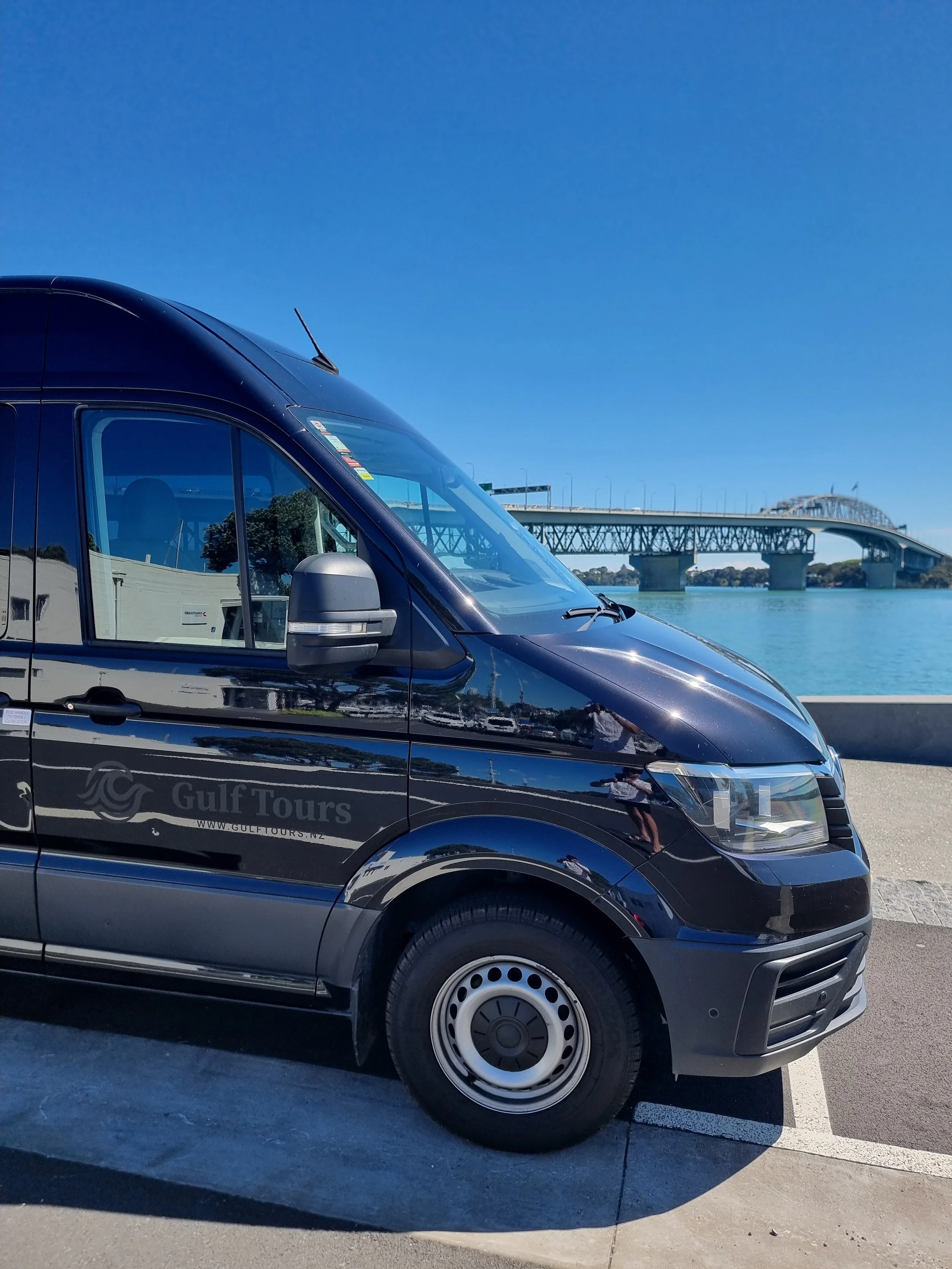 Black van with Gulf Tours logo parked near water with the Auckland Harbour bridge and the Waitemata Harbour in background under clear blue sky.