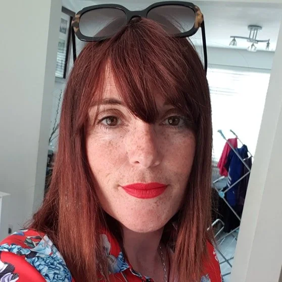 A woman with red hair, freckles, and red lipstick is taking a selfie. She is wearing sunglasses on her head and a patterned red blouse. In the background, there is a laundry drying rack with clothes hanging on it.