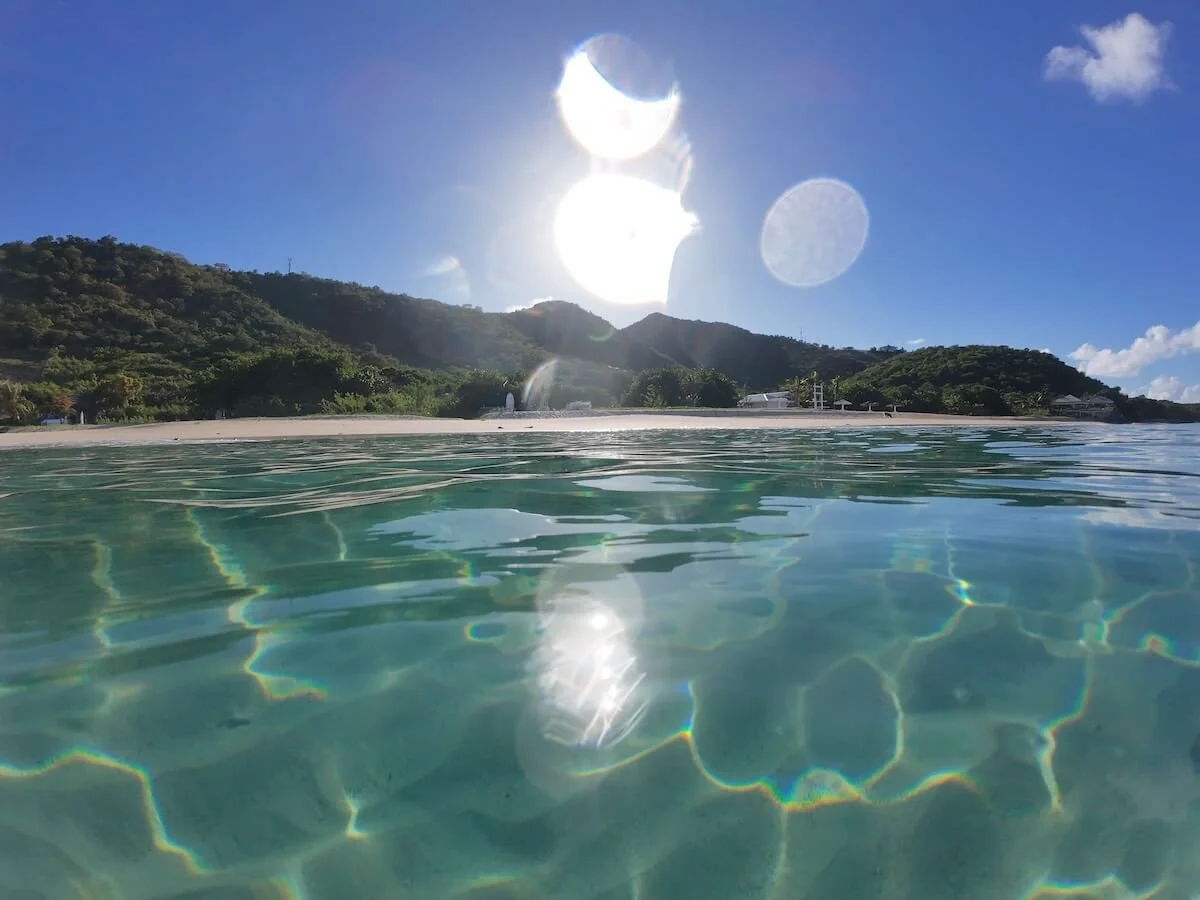 Overwater view of a Crabbe Hill beach in Antigua with clear turquoise waters, green hills, and the sun making sun spots in the sky and water, the destination for The Sea Change Retreat to receive the clarity and inspiration for your New Era.