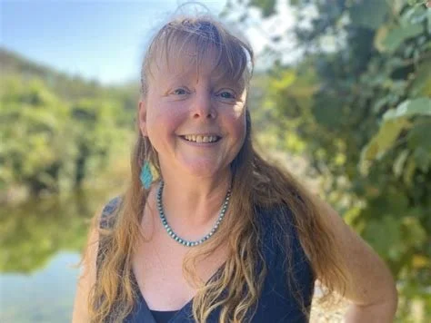 Katie Sarra, a former client of Kat Byles and advocate of the True Business model smiling standing outdoors near a body of water and trees, inspired to live in harmony with nature, ourselves and each other