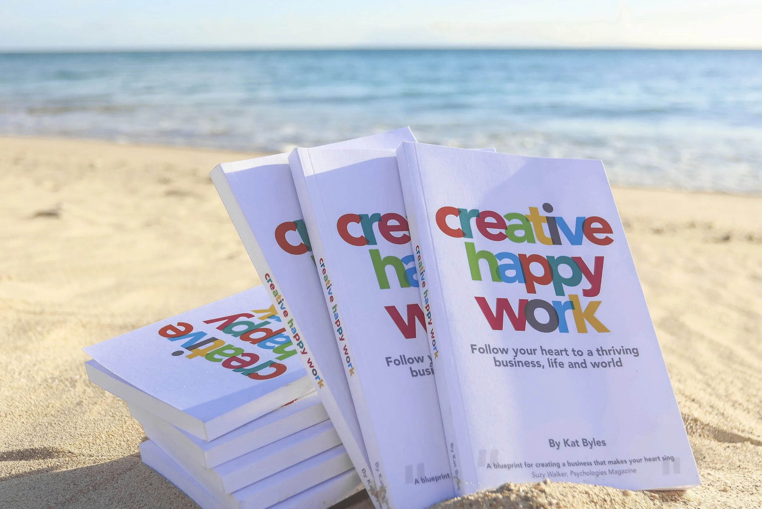 Multiple copies of the book 'Create Happy Work' arranged on a sandy beach with the ocean and blue sky in the background.