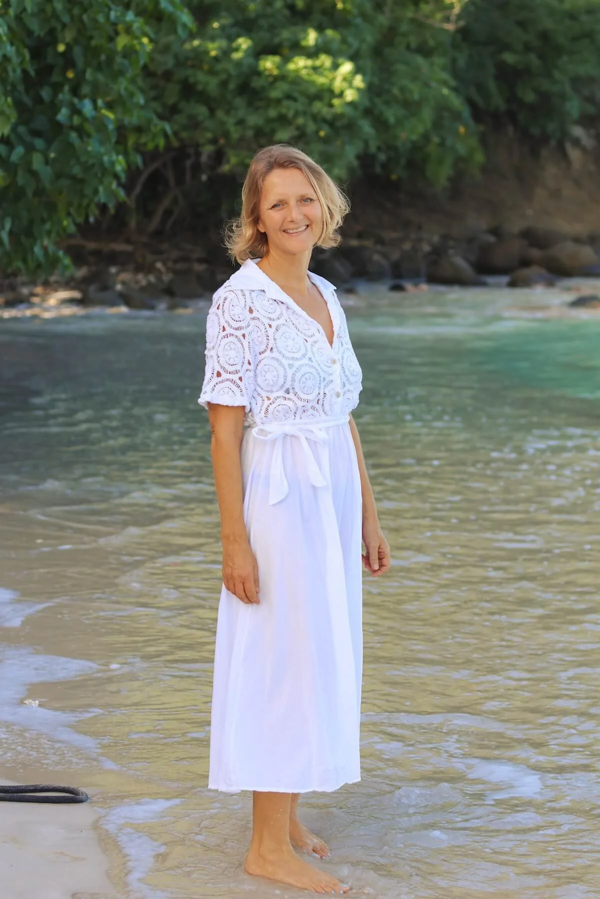 Kat Byles, author Creative Happy Work and host of The Sea Change Retreat in Antigua is standing barefoot on the sandy shore of a beach in Antigua, smiling, with green trees in the background and the ocean behind her.
