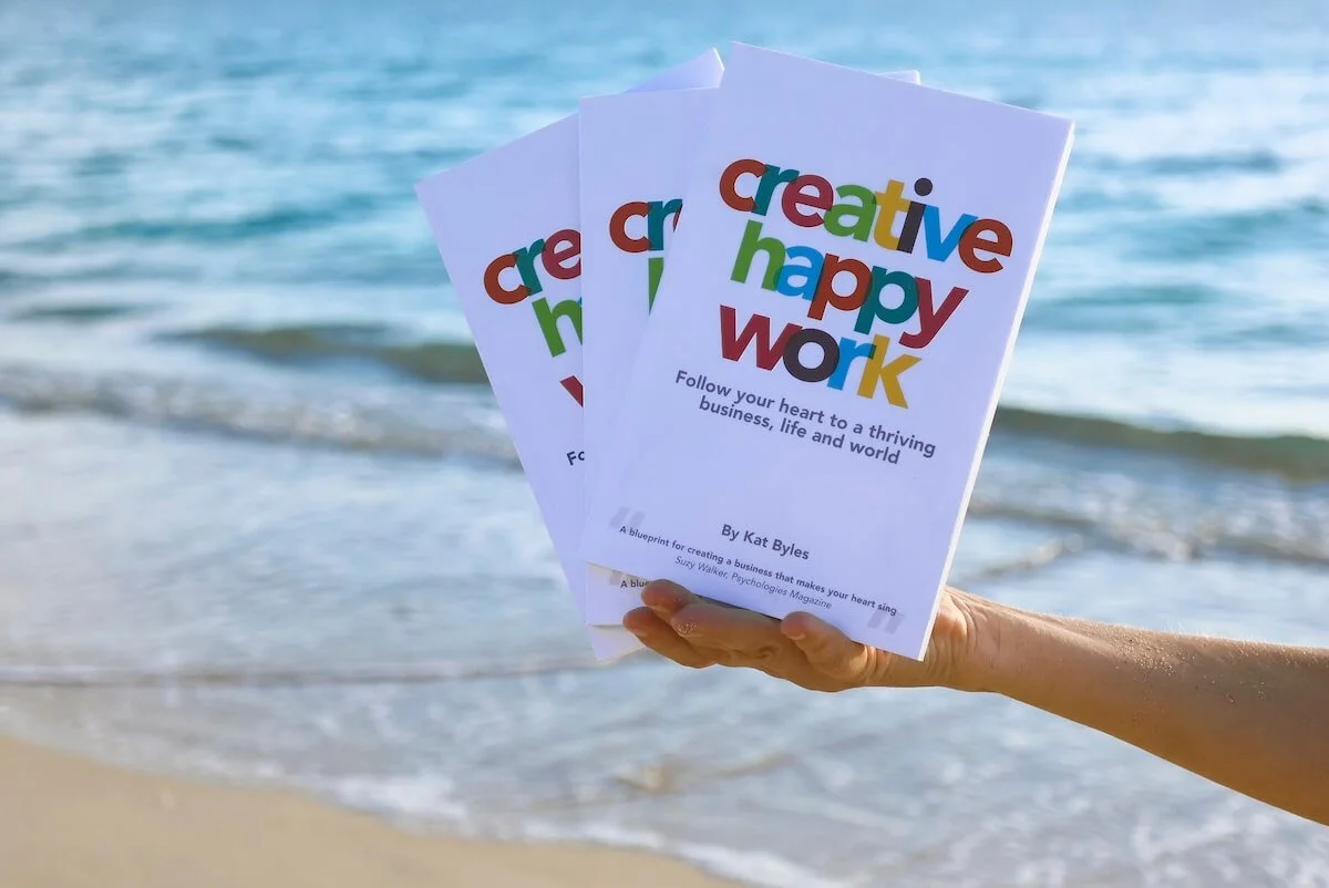 Kat Byles, author of Creative Happy Work, holds out her hand with three copies of Creative Happy Work against a Caribbean Sea backdrop, the inspiration for the True Business model to build your business with alignment not hustle