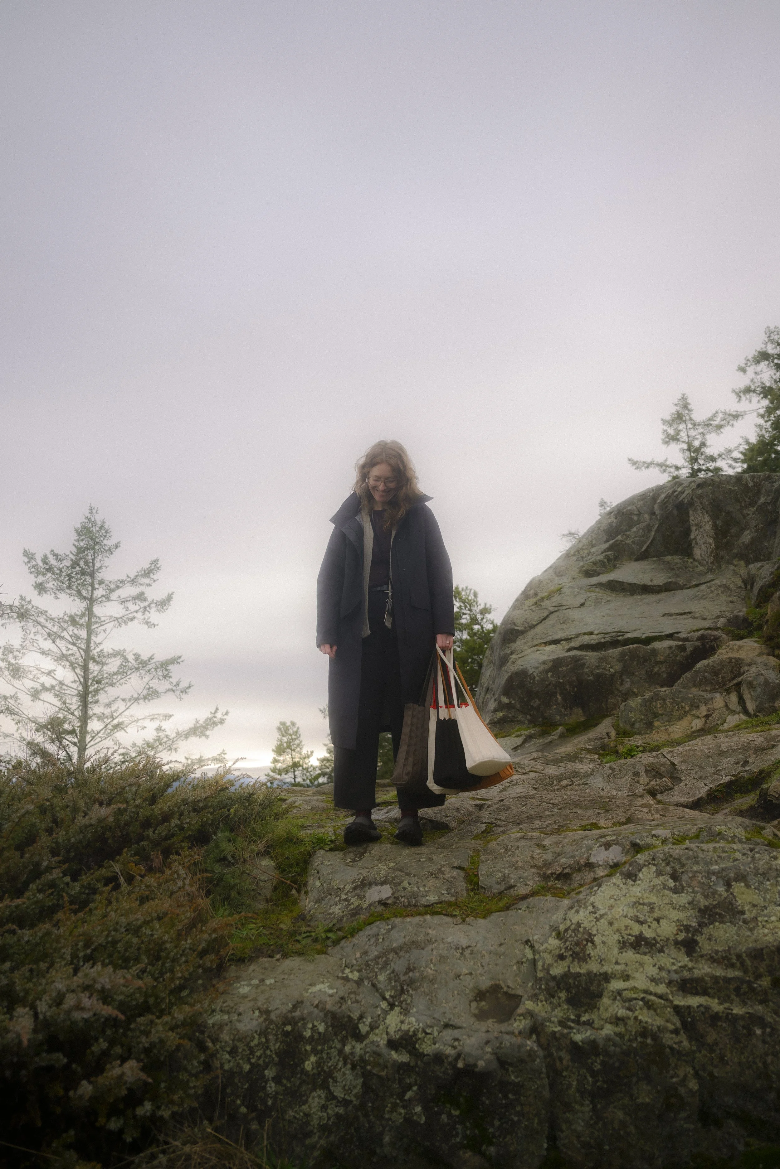 Woman in black coat and glasses smiling while standing on rocky terrain outdoors, holding shopping bags, with trees and overcast sky in the background.