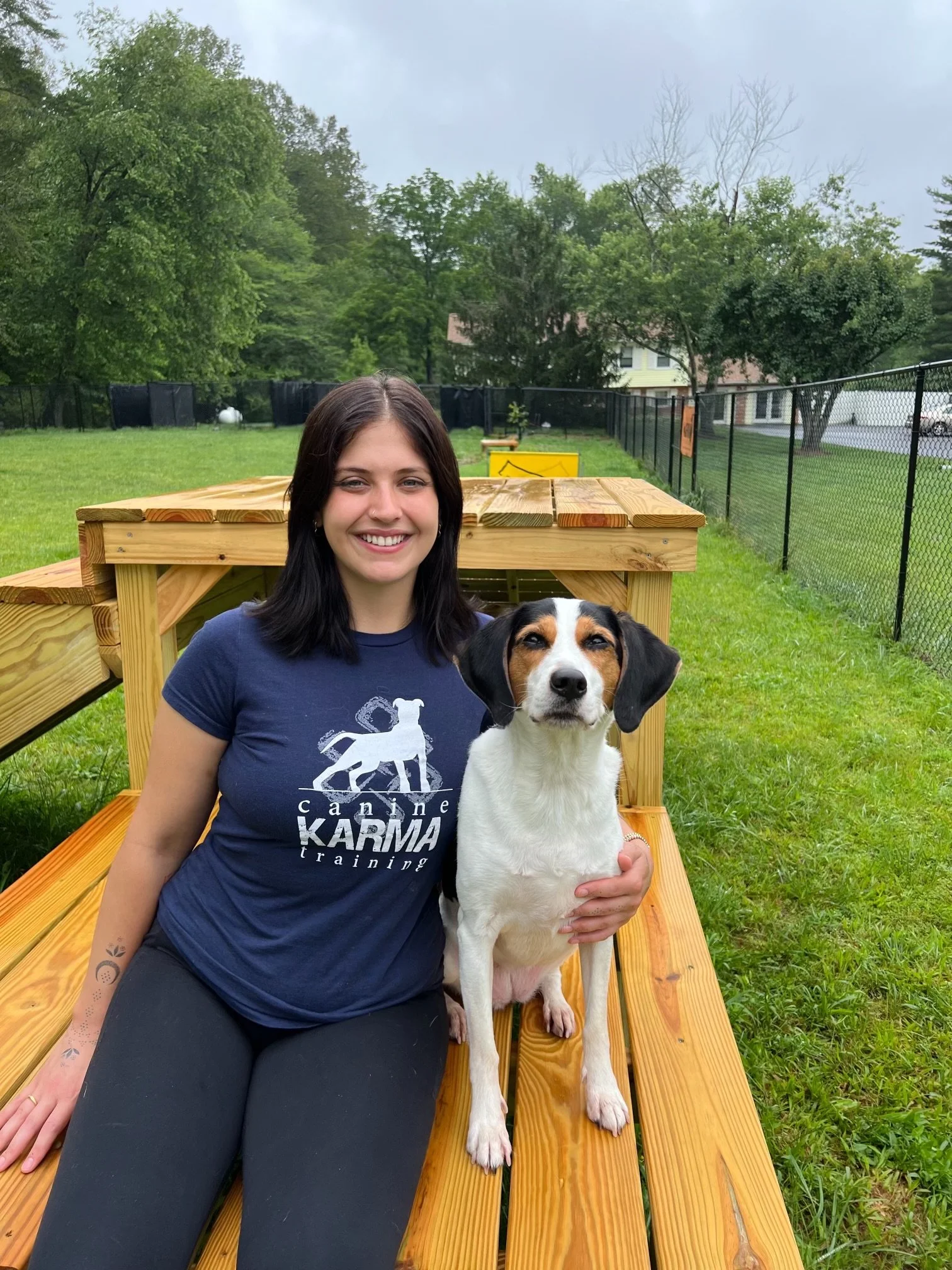 A woman sitting on a wooden platform outdoors, smiling, with a dog sitting beside her, in a grassy backyard with trees and a chain-link fence.