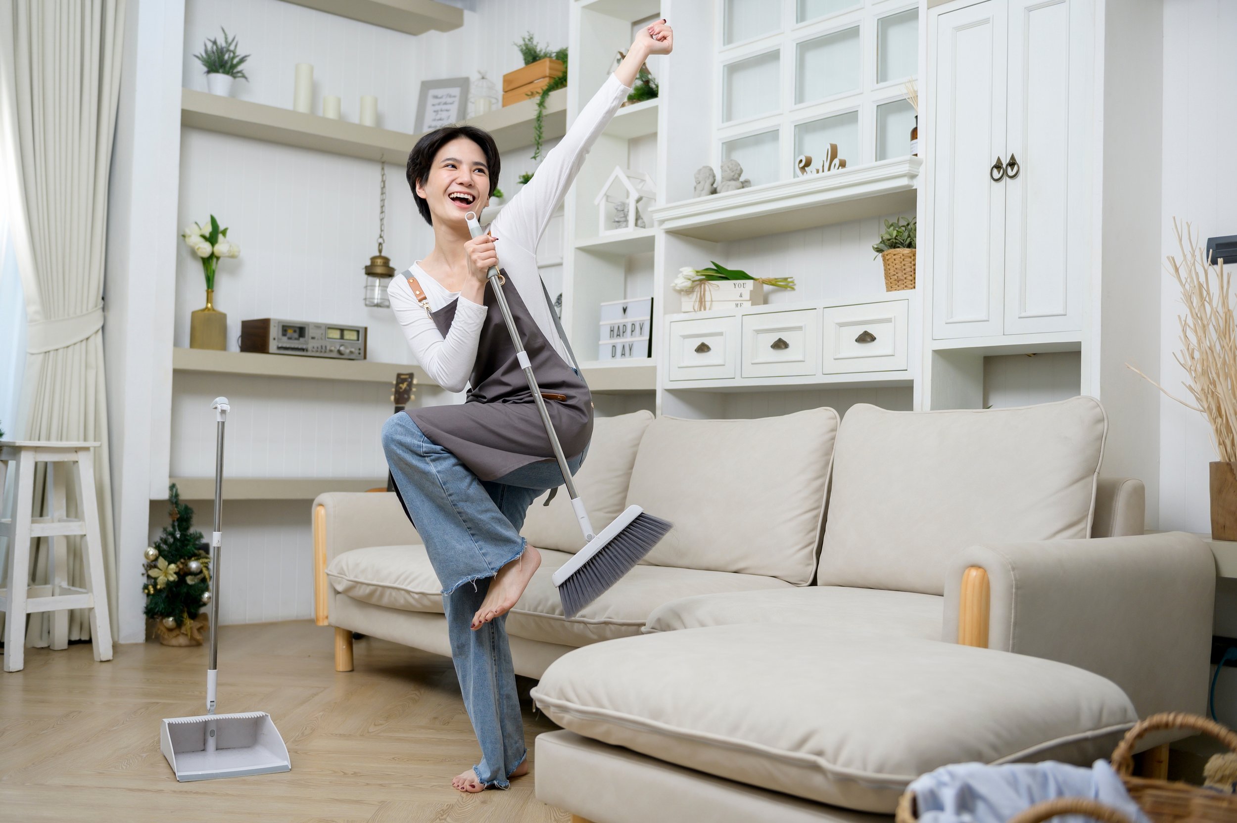 A woman cheerfully singing while holding a mop in a bright living room, with one foot on a sofa, and surrounded by cleaning tools and decorative items.