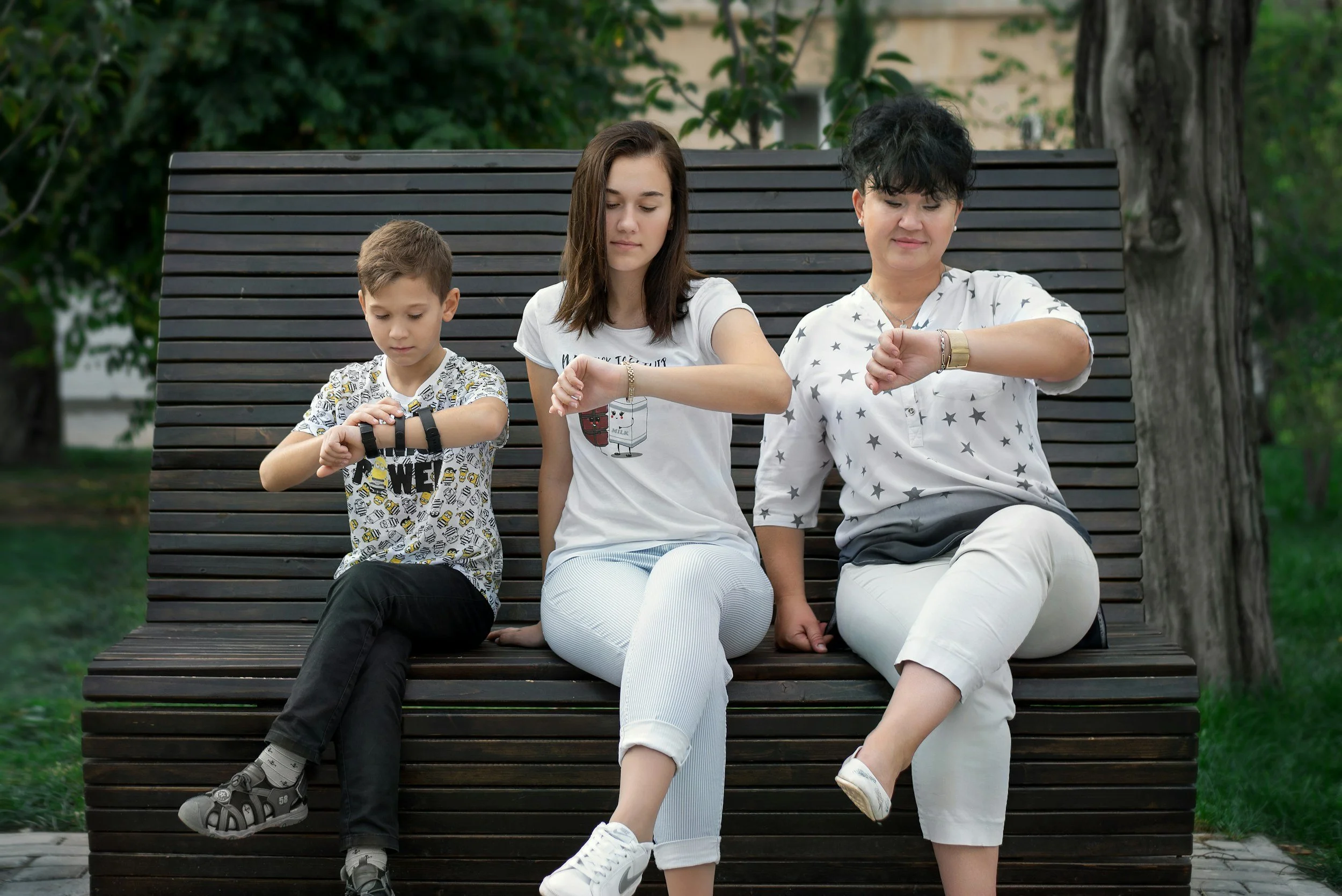 A woman with dark, curly hair and a white shirt with black stars, a teenage girl with straight brown hair and a white t-shirt, and a young boy with short brown hair and a patterned shirt, sitting on a wooden park bench outdoors, looking at their watches.