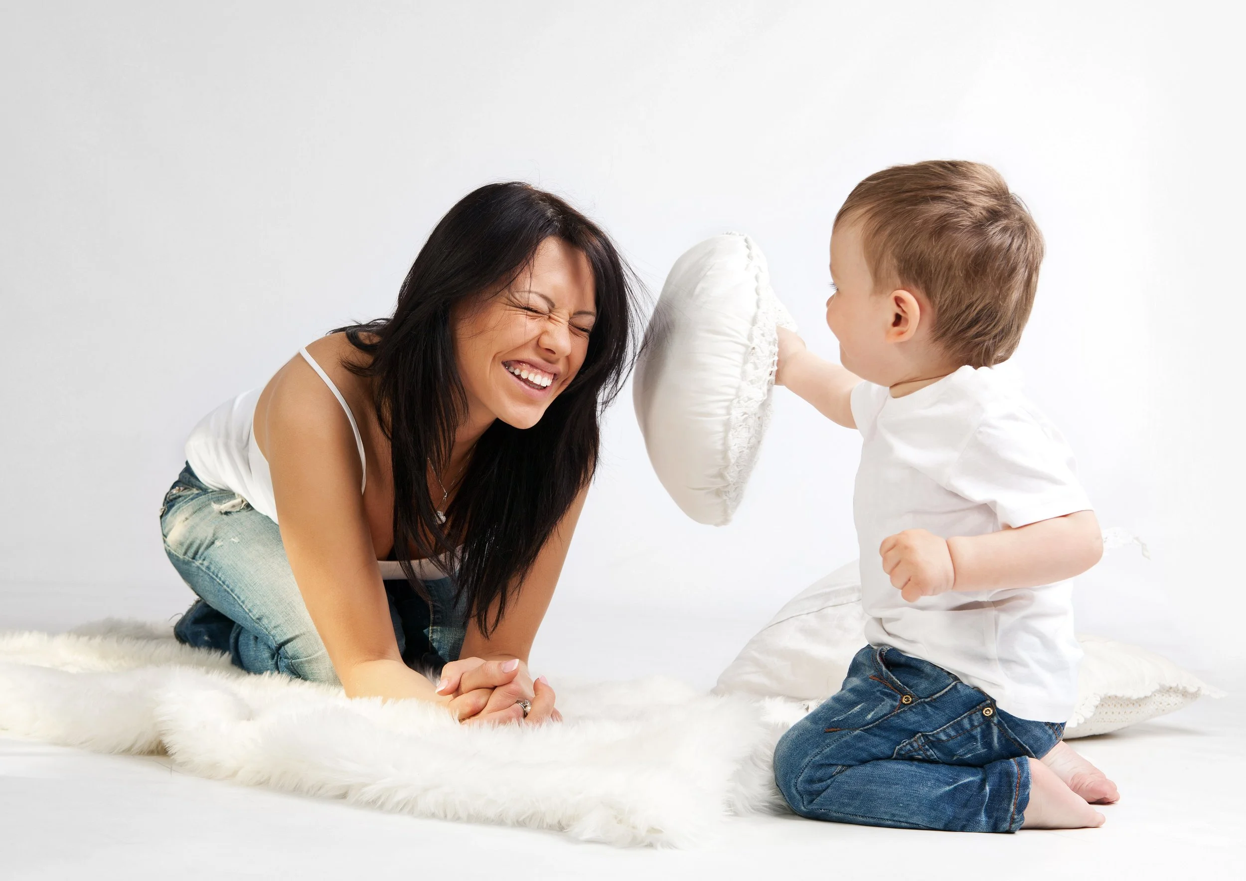 A woman and a young boy playing gently with a pillow on a white furry rug in a bright room, both smiling and enjoying their time together.