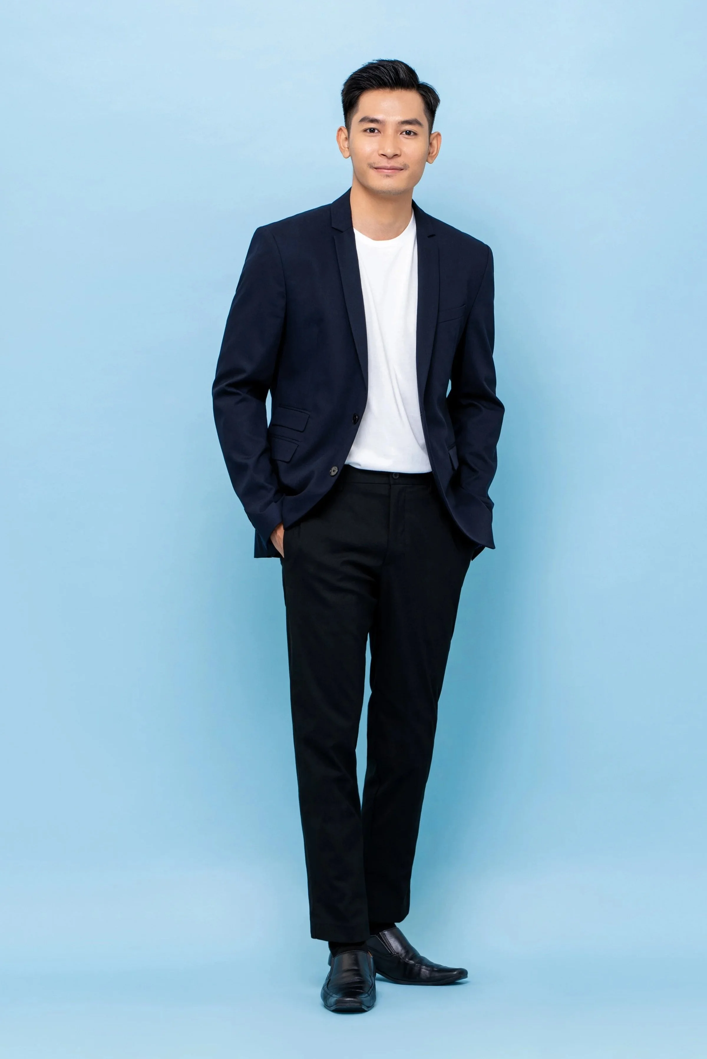 A young man in a navy blue blazer, white t-shirt, black dress pants, and black shoes standing against a light blue background with a confident smile.