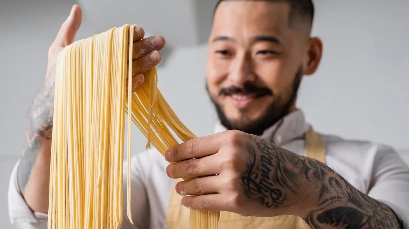 A male chef with a shaved head and a stubble beard, wearing a white chef's coat and a black apron, standing with his arms crossed in a professional kitchen.