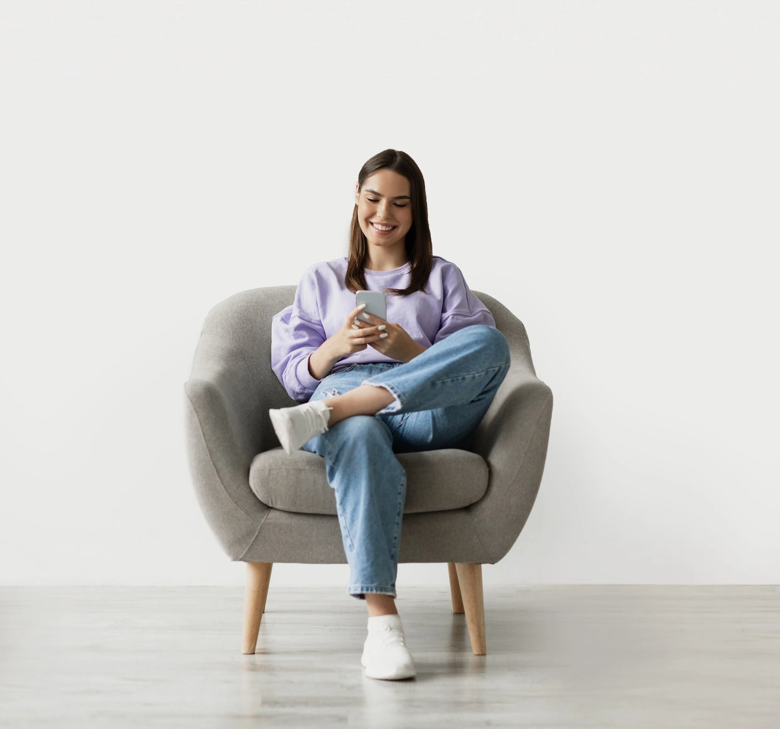 Smiling woman in cozy armchair checking her phone — representing the comfort and confidence of households supported by GoodMaids.