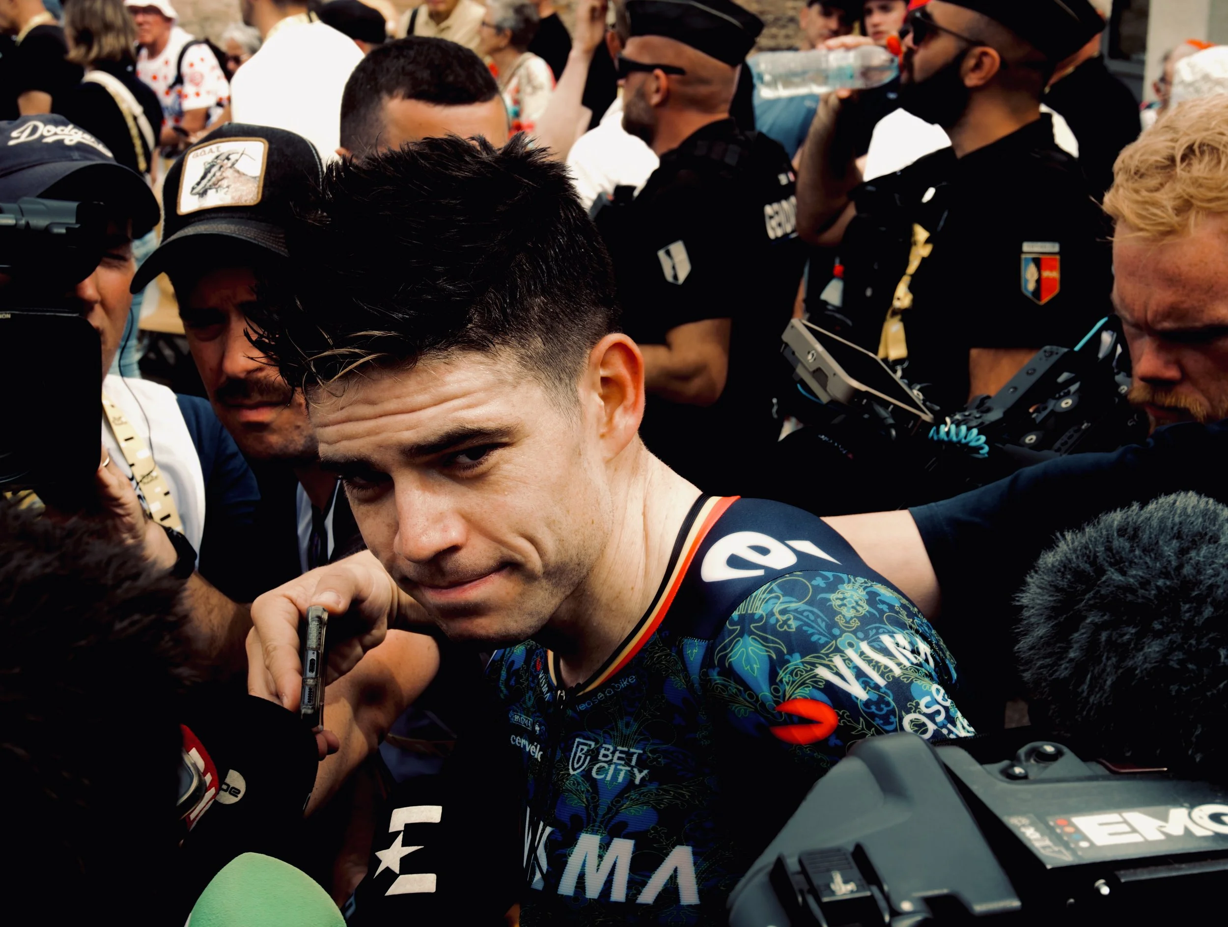 Un homme, probablement un cycliste, est entouré de médias et de spectateurs, alors qu'il semble répondre aux questions après une course ou un événement public.