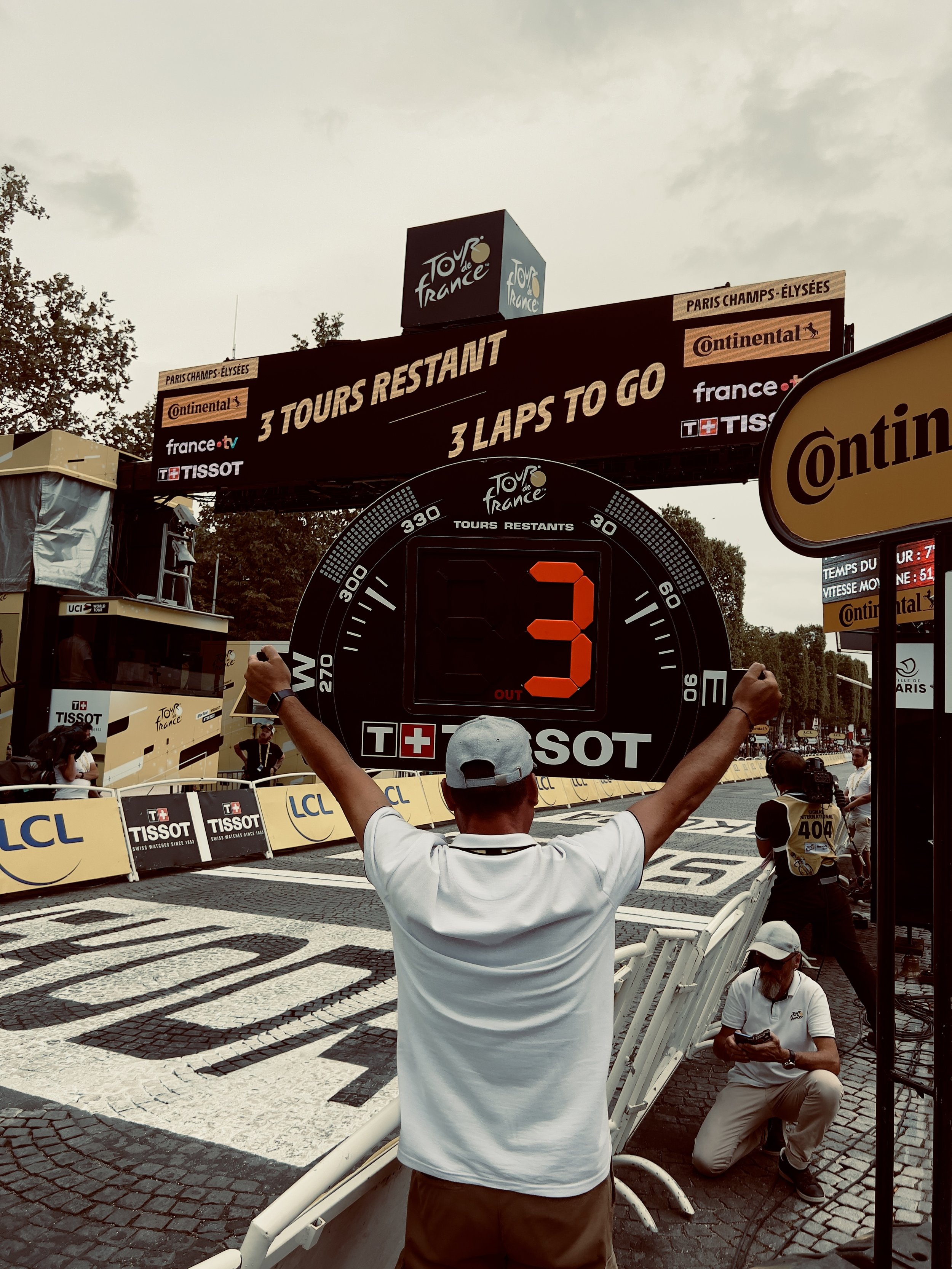 Une personne en tenue blanche avec une casquette blanche, levant les bras devant un tableau numérique affichant le nombre 3, lors d'une étape du Tour de France. Il y a une autre personne en arrière-plan, assise et utilisant un téléphone, et un pannea