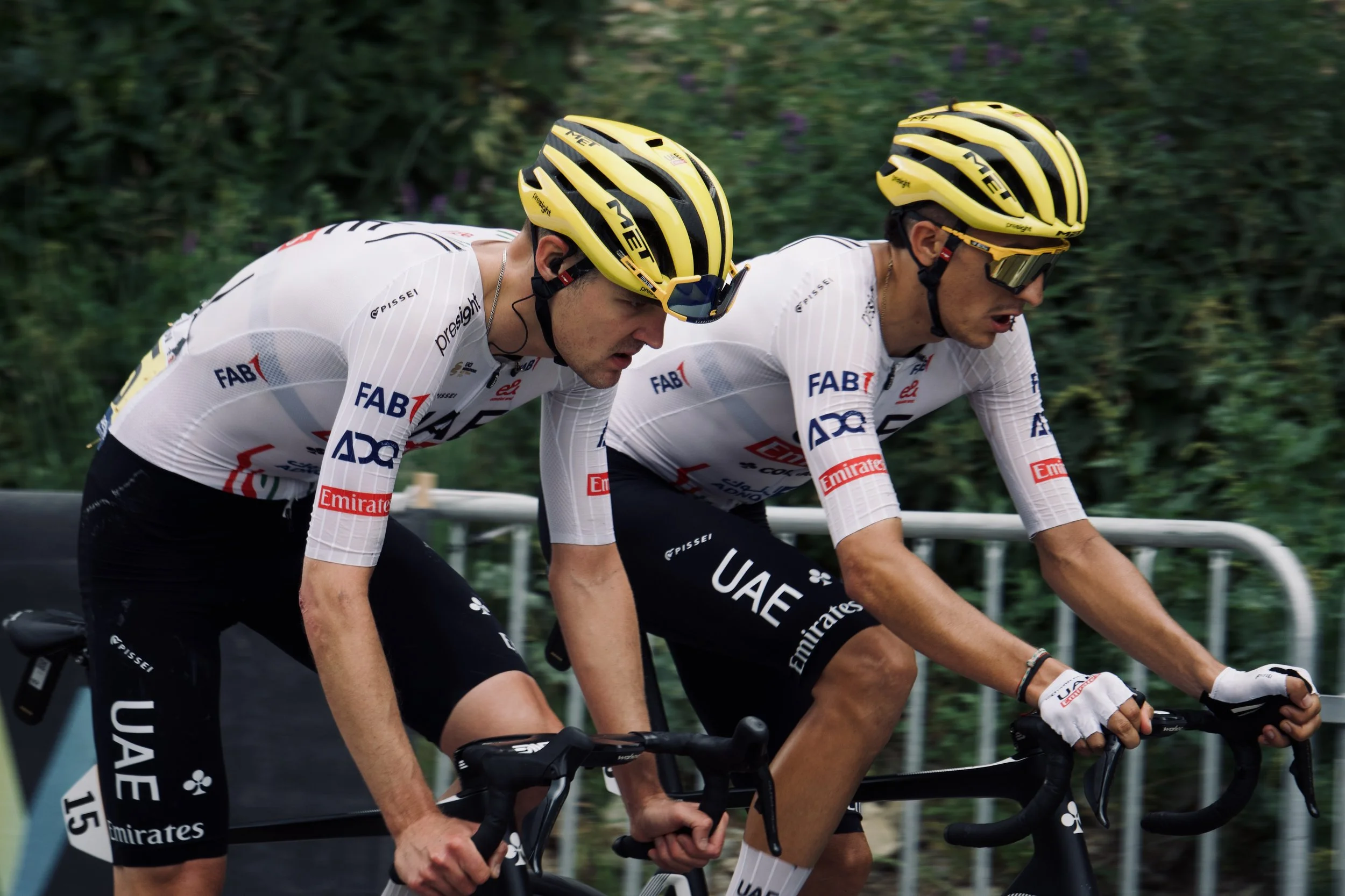 Deux cyclistes en course portant des maillots de l'équipe UAE, casque jaune, lunettes noires, pédalant rapidement.