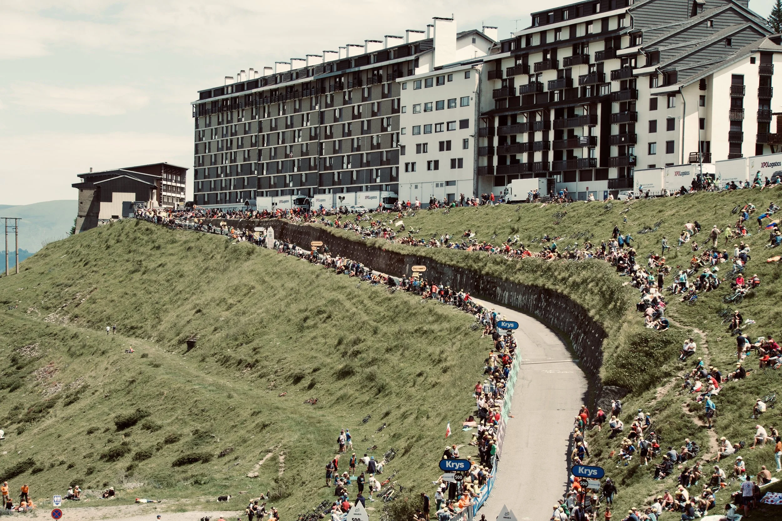 Une foule de spectateurs assis ou debout le long d'une route escarpée entourée de collines verdoyantes, avec des immeubles résidentiels modernes en arrière-plan, lors d'un événement en plein air.