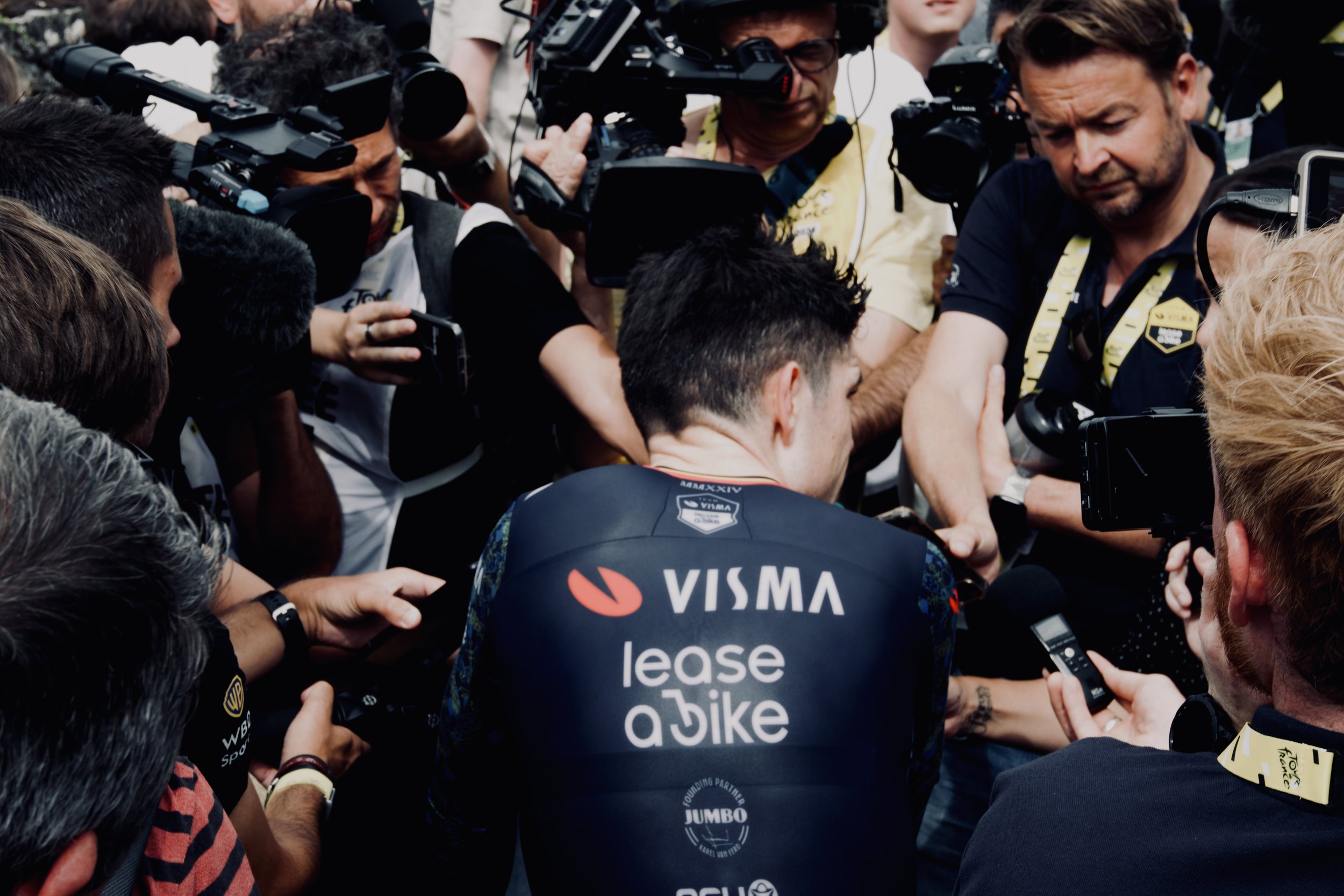Un coureur cycliste entouré par des journalistes et des caméramans, portant un maillot avec des logos "VISMA" et "lease a bike".
