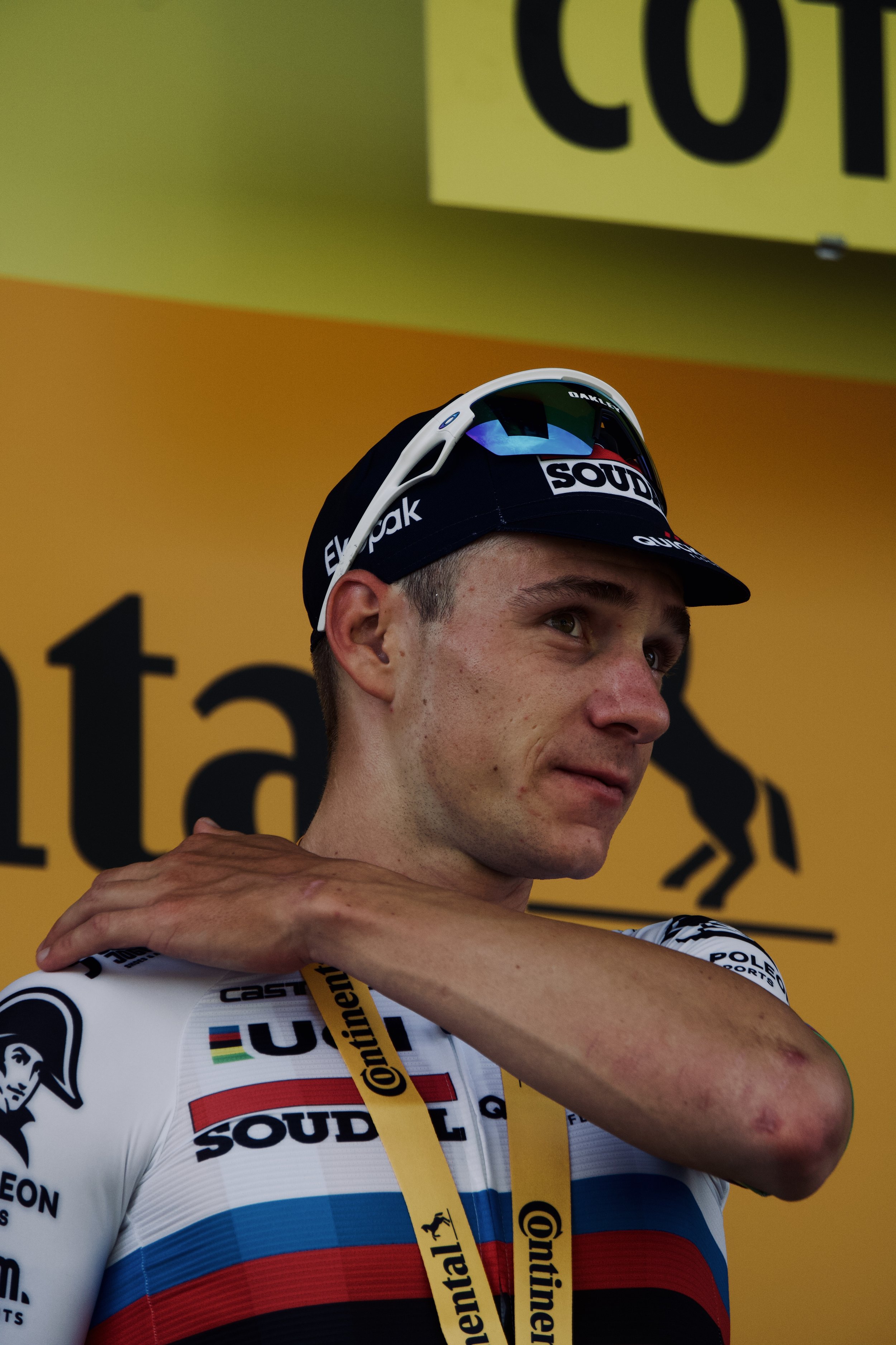 Un cycliste en tenue de course, portant une médaille, se tenant près d'un fond jaune avec des textes noirs, après une compétition.