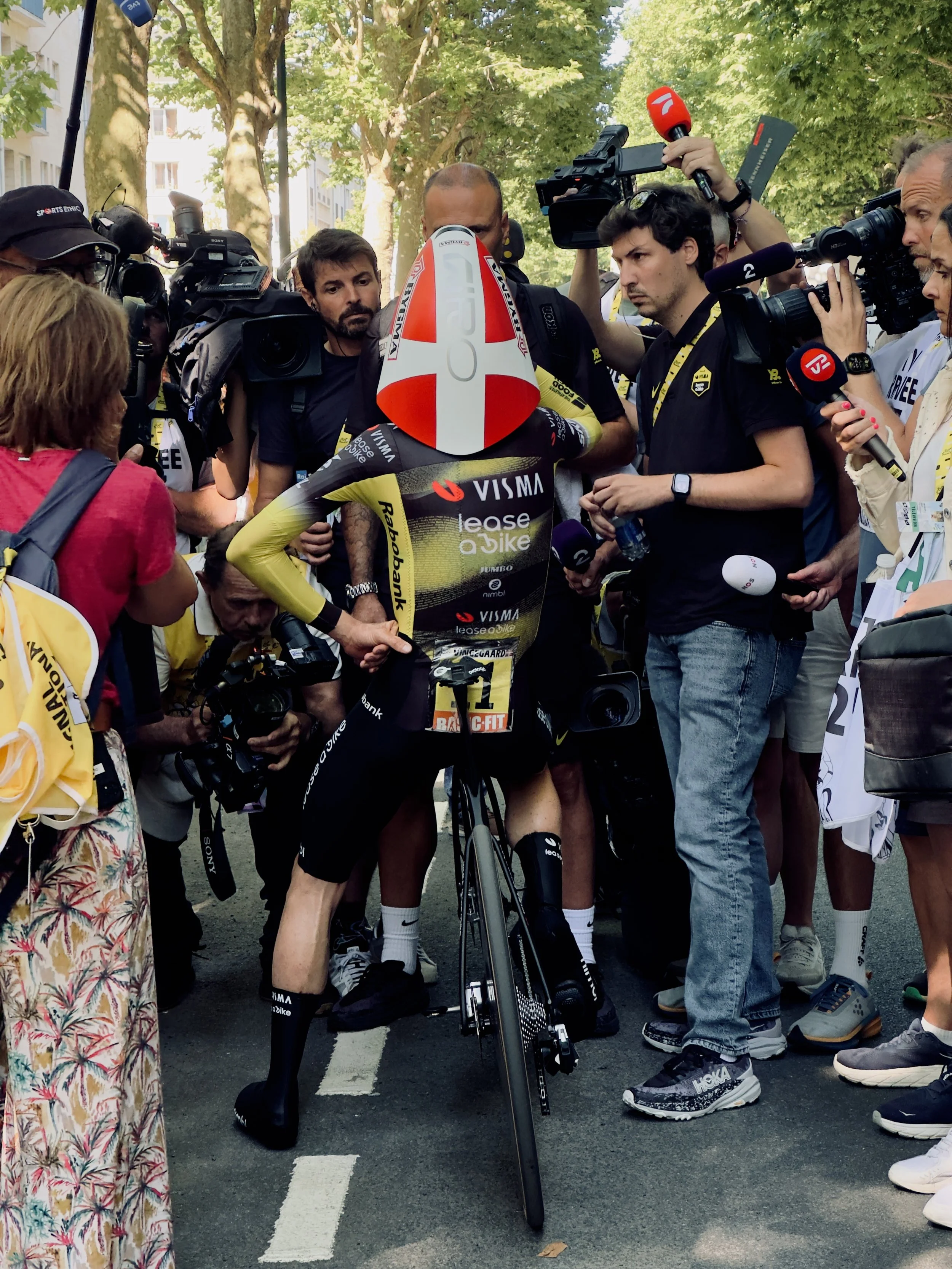 Un cycliste sponsorisé par Visma Lease vient de terminer une course, entouré de journalistes, photographes et spectateurs dans une zone urbaine ombragée.