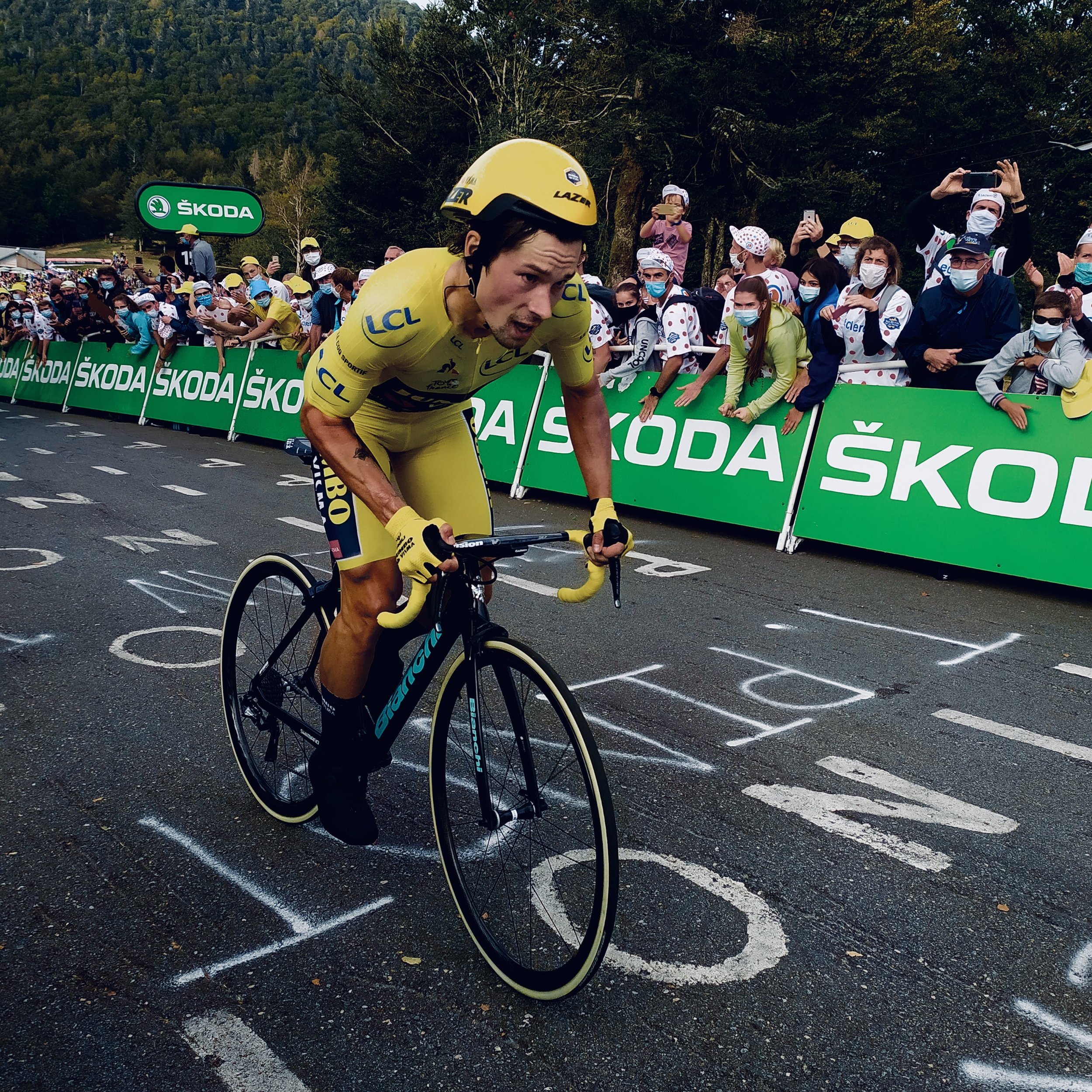 Un cycliste en maillot jaune franchit la ligne d'arrivée lors d'une compétition, avec des spectateurs en arrière-plantenant des téléphones pour prendre des photos, entourés de barrières publicitaires vertes.