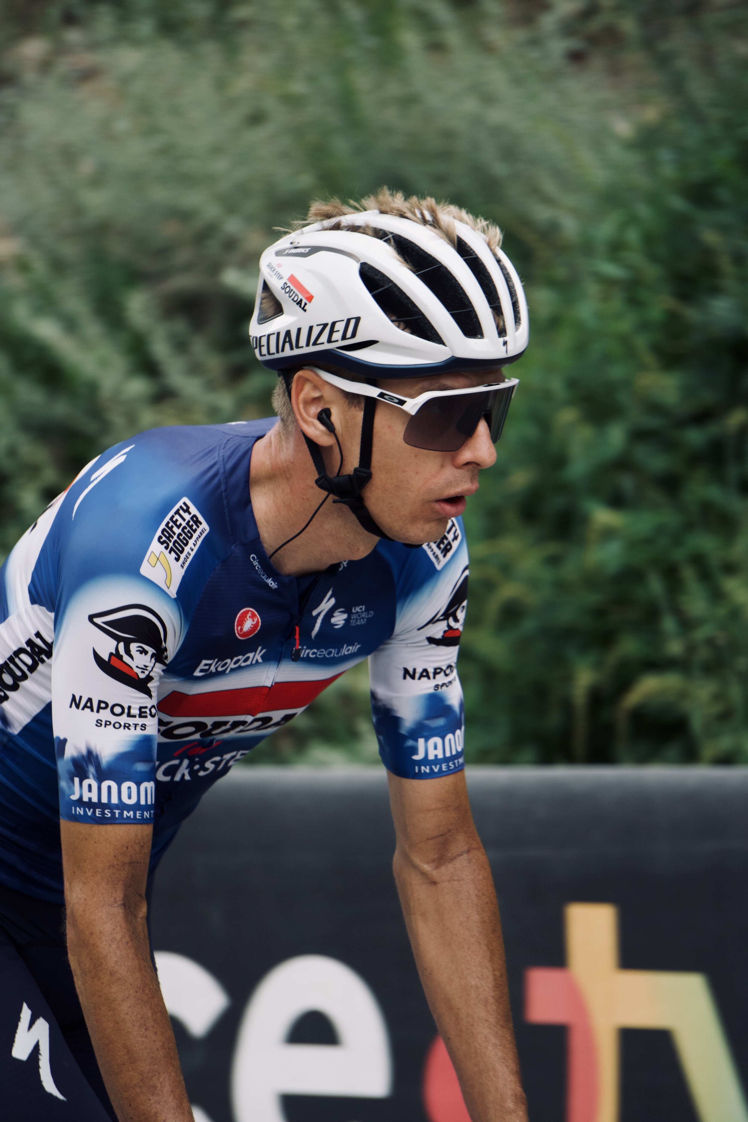 Un cycliste portant un casque blanc et des lunettes noires, vêtu d'un maillot de cyclisme bleu et blanc avec des logos, en train de rouler dans un environnement naturel.