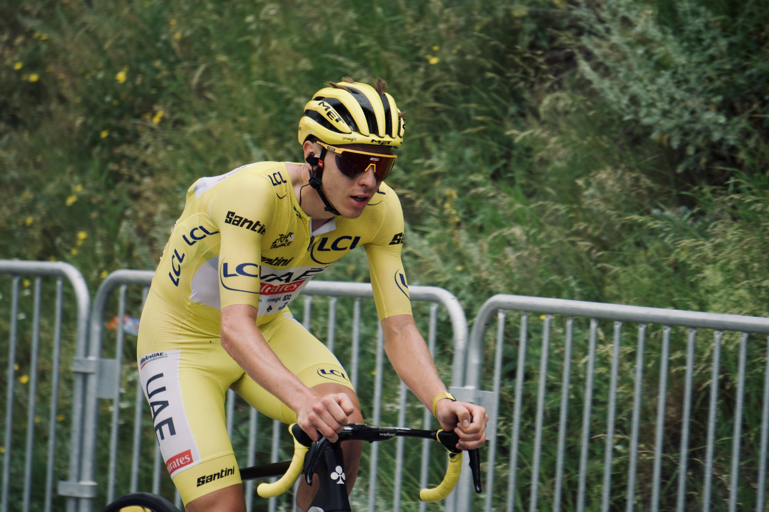 Un cycliste en tenue jaune portant un casque et des lunettes de soleil, en pleine course sur route, avec des barrières en métal en arrière-plan.