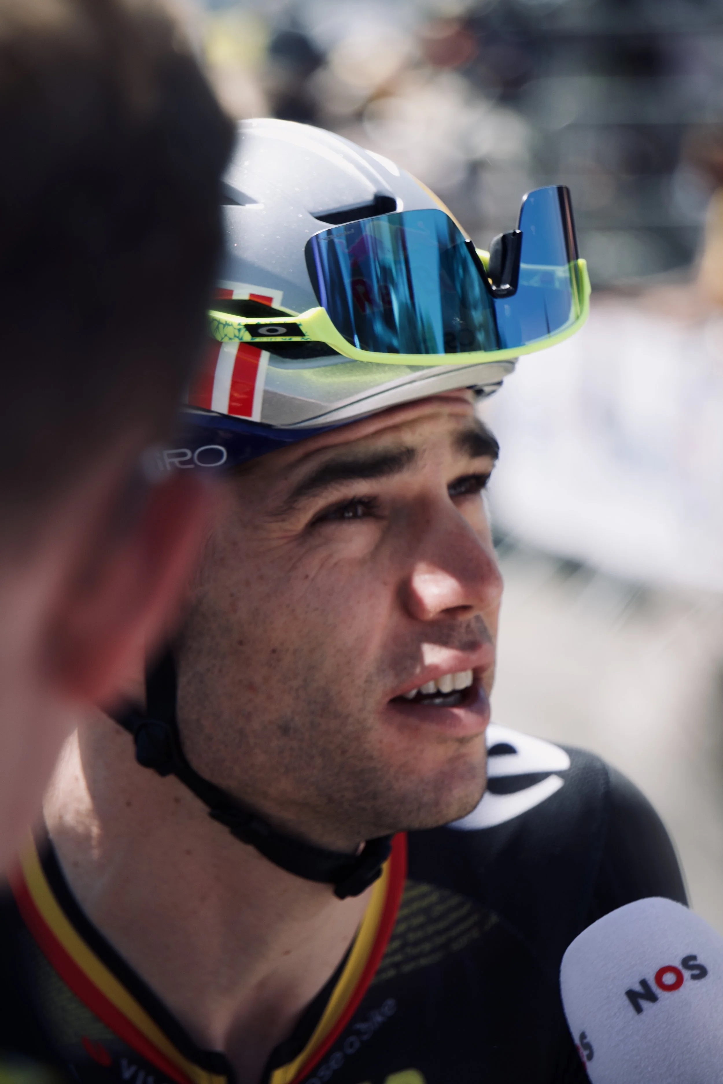 Un homme avec un casque de cyclisme et des lunettes de soleil, parlant à une interview, portant un maillot noir avec des accents jaunes et rouges.