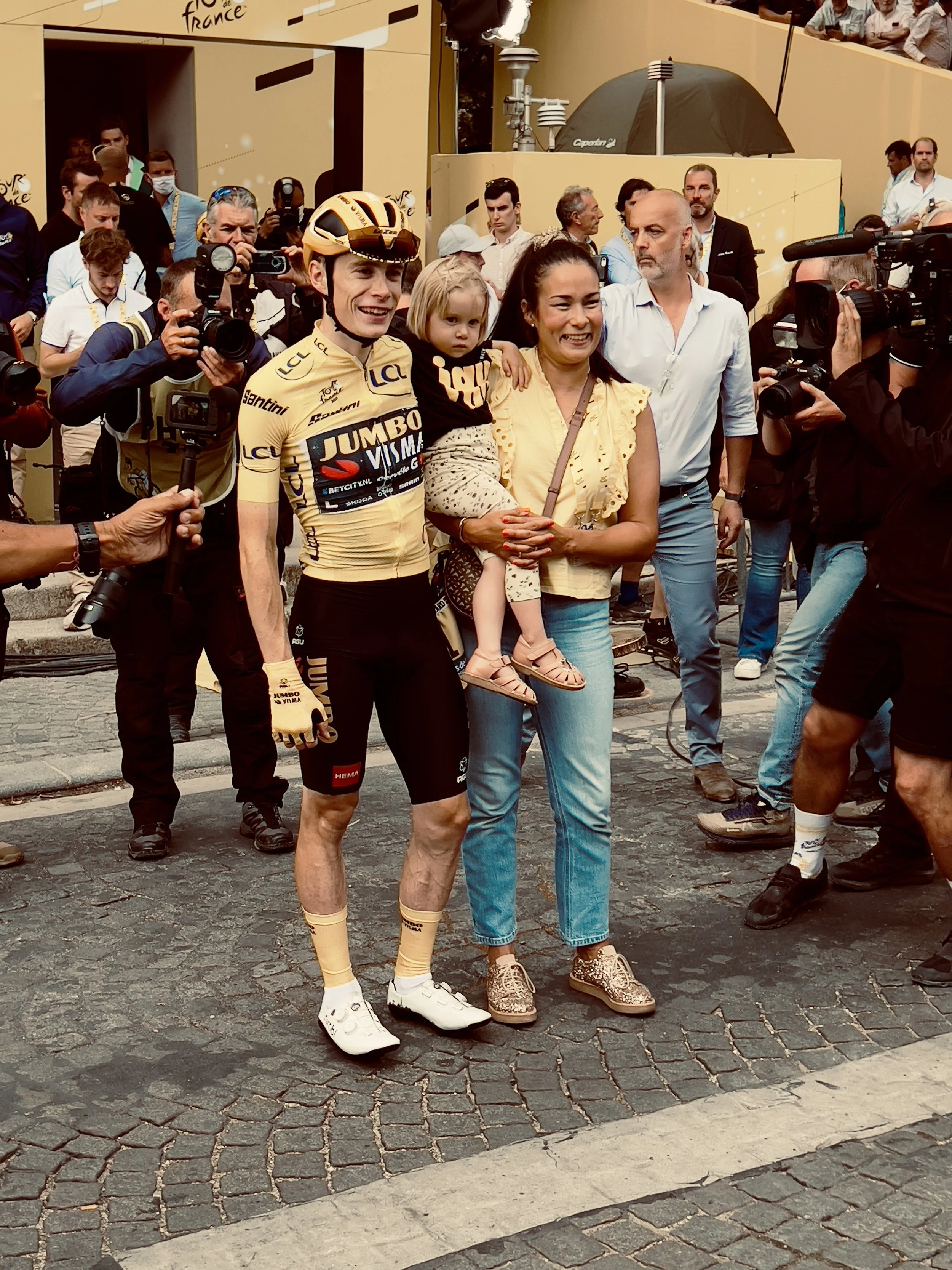 Un cycliste en uniforme jaune de la Jumbo-Visma posant avec une famille après une compétition, entouré de journalistes et de caméras.