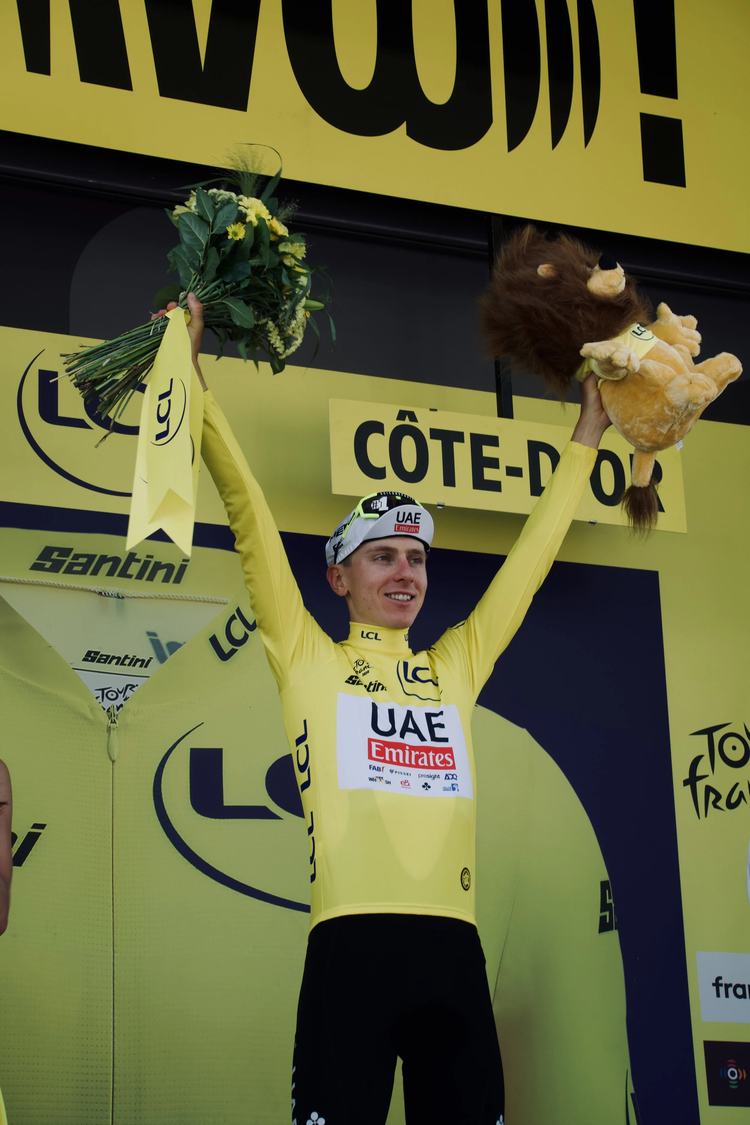 Un coureur cycliste en maillot jaune, debout sur le podium avec les bras levés. Il tient un bouquet de fleurs dans une main et un lion en peluche dans l'autre, célébrant sa victoire.
