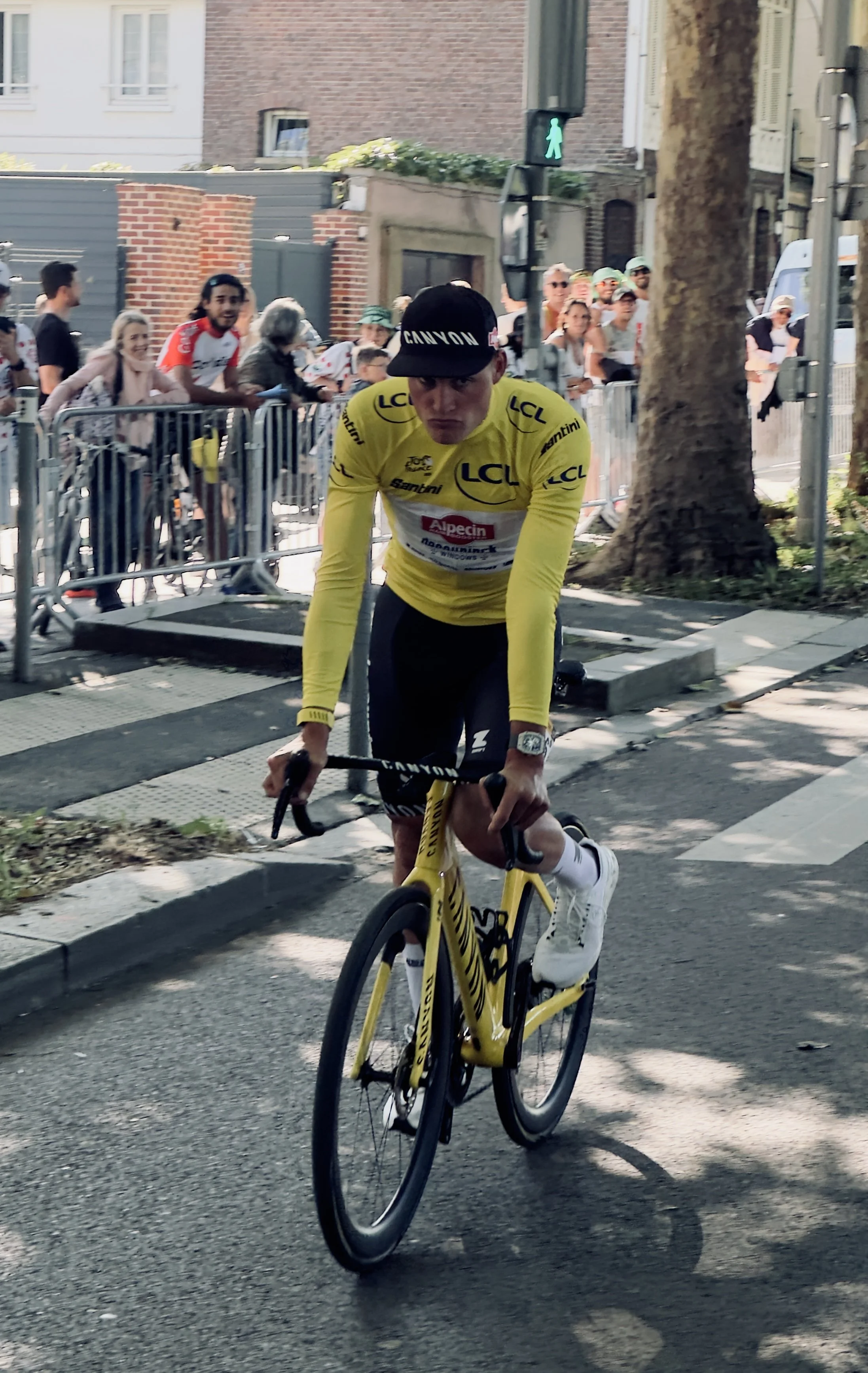 Un cycliste portant un maillot jaune et un chapeau noir, en train de rouler sur la route lors d'une course, avec des spectateurs derrière des barrières en train de l'encourager.