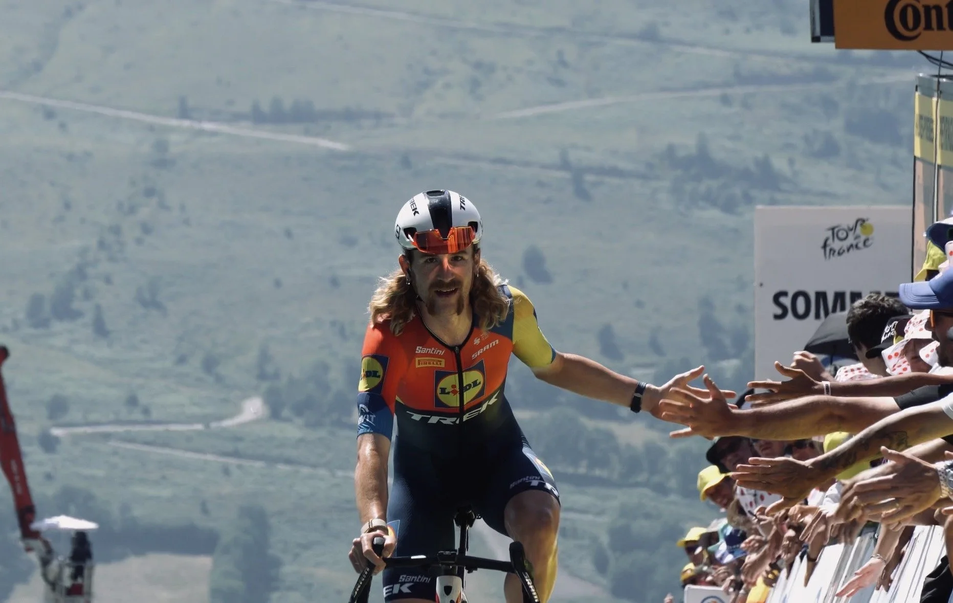 Un cycliste en tenue de compétition, saluant des spectateurs lors du Tour de France, avec une foule applaudissant derrière une barrière sur un fond de paysage montagneux.