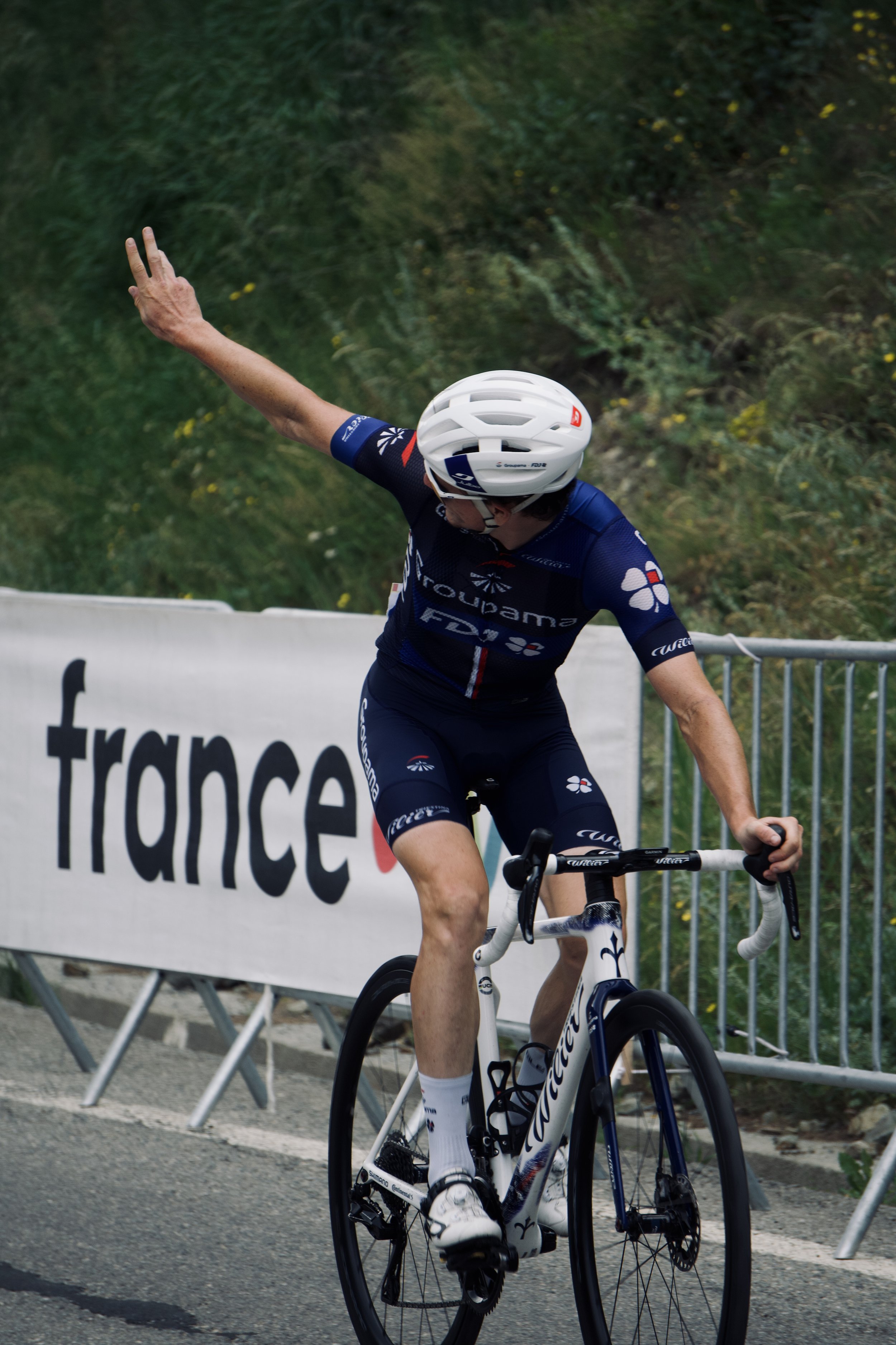 Un cycliste portant un maillot bleu de l'équipe Groupama-FDJ, casque blanc, en train de faire la course sur une route. Il semble célébrer ou saluer, avec une bras levé.