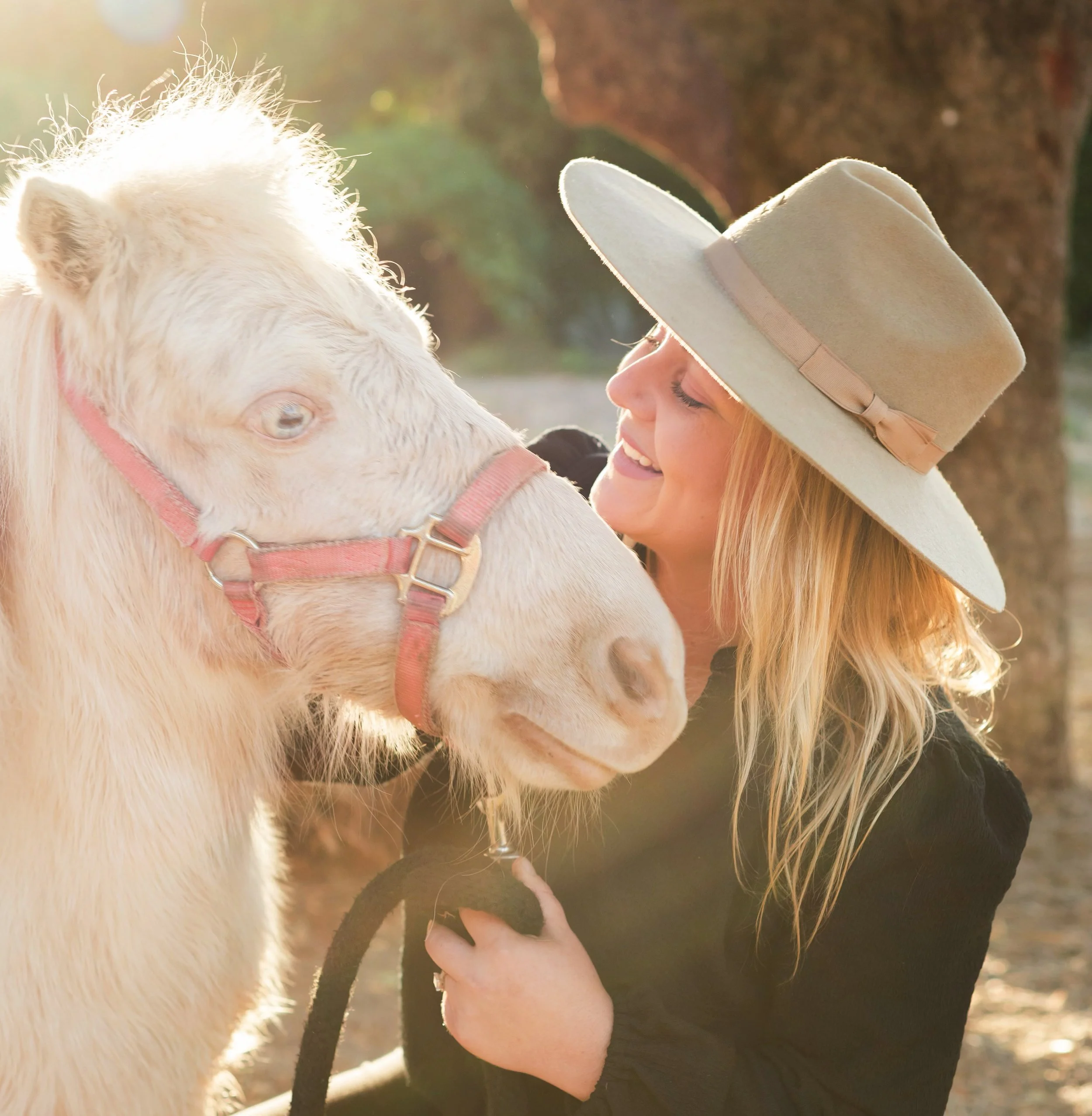 A woman with long blonde hair wearing a large beige hat and black shirt smiling and holding a white horse with a pink halter outdoors near a tree.