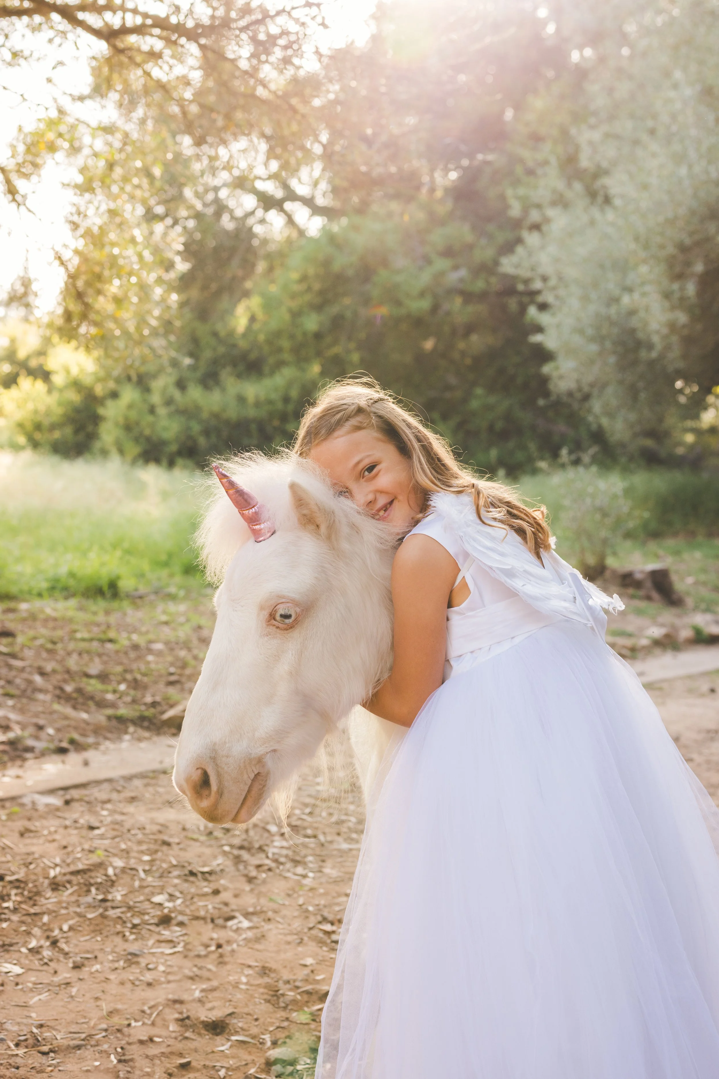 A young girl in a white dress hugging a white unicorn with a pink horn in a sunlit park.