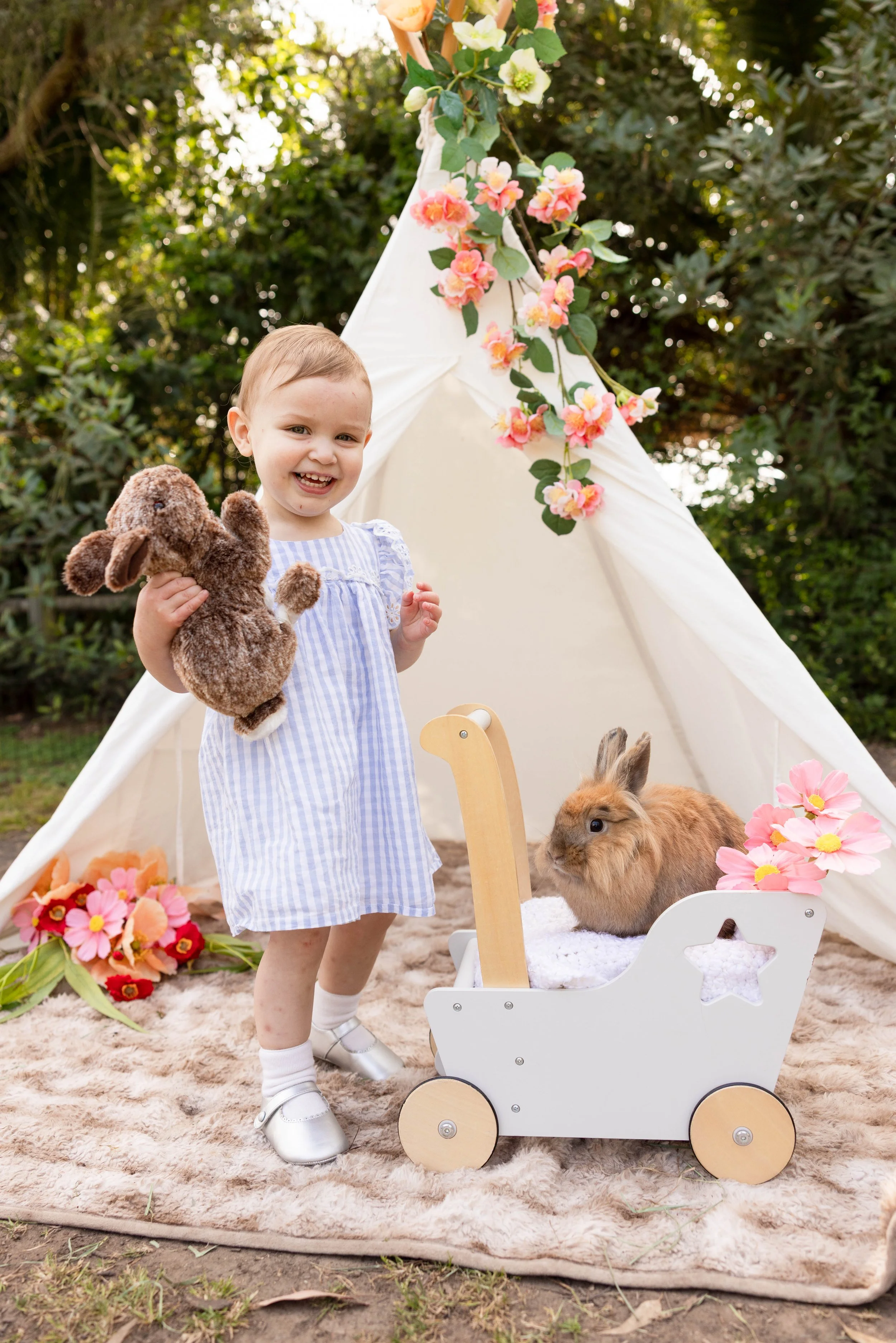 A young girl with short hair smiling and holding a stuffed animal bunny in front of a white decorative stroller with a rabbit inside. Behind her, there is a white teepee decorated with pink and green flowers on a green backyard setting with trees.