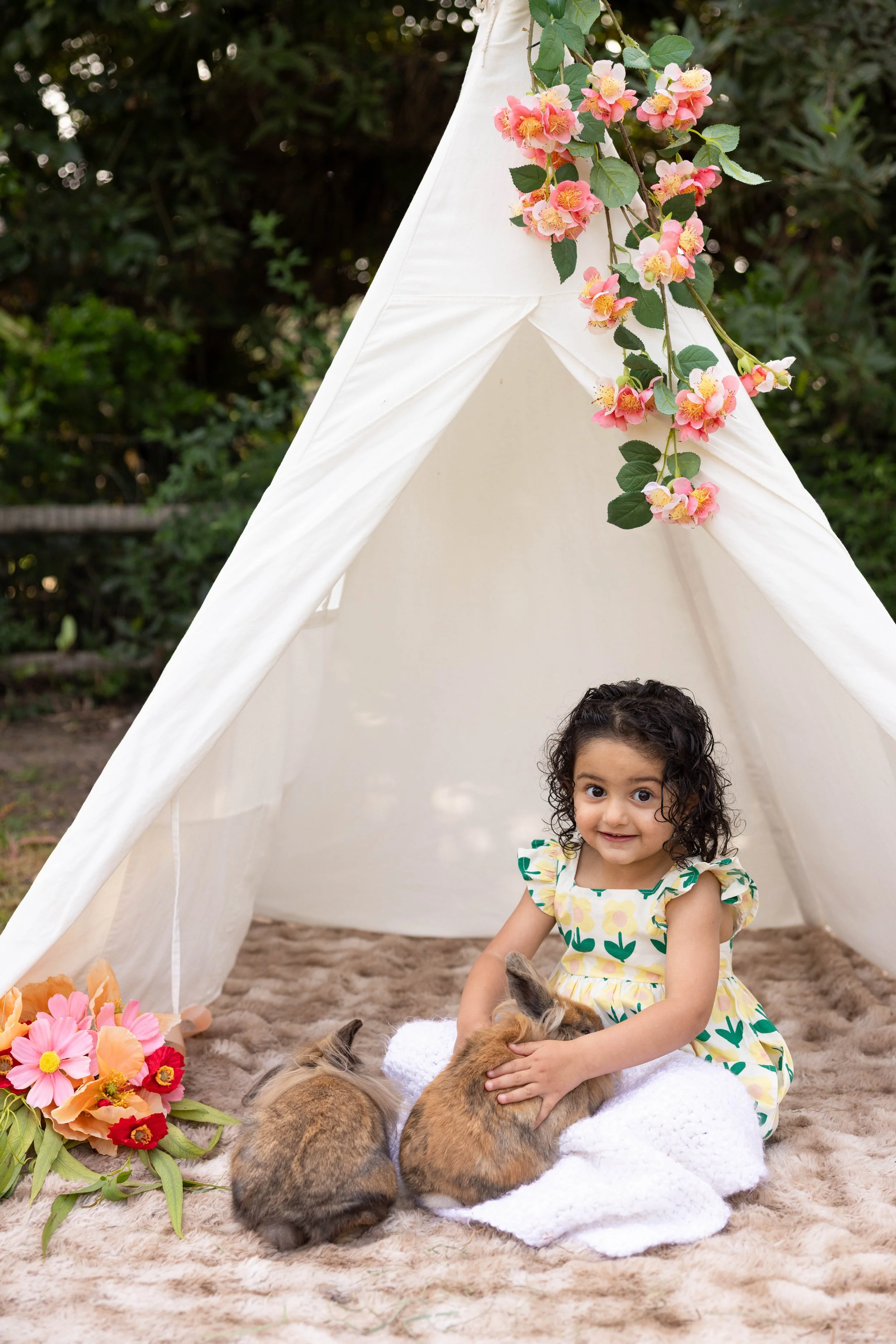 A young girl with curly dark hair, wearing a colorful dress with green and yellow patterns, sitting on a beige plush rug inside a white tent with pink and orange flower decorations hanging above. She is petting a small brown and black rabbit with two