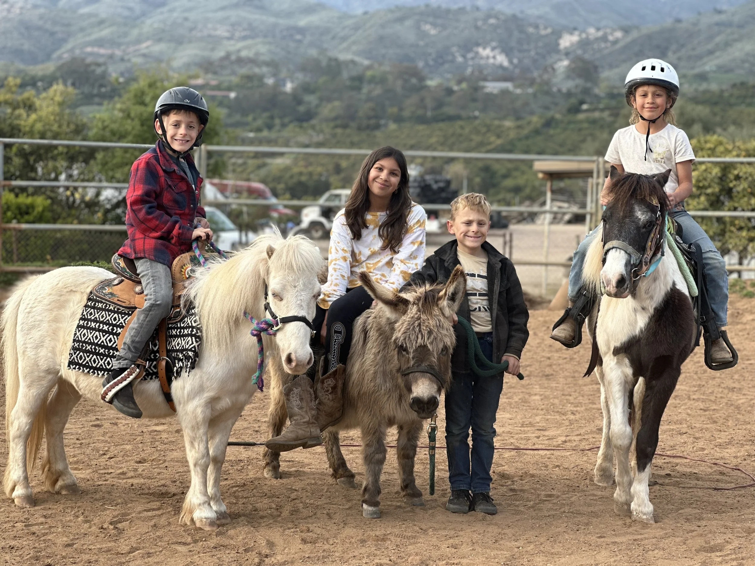 Four children with three ponies in a dirt riding arena, with a fence and hills in the background. Two children are riding ponies, while two are standing.