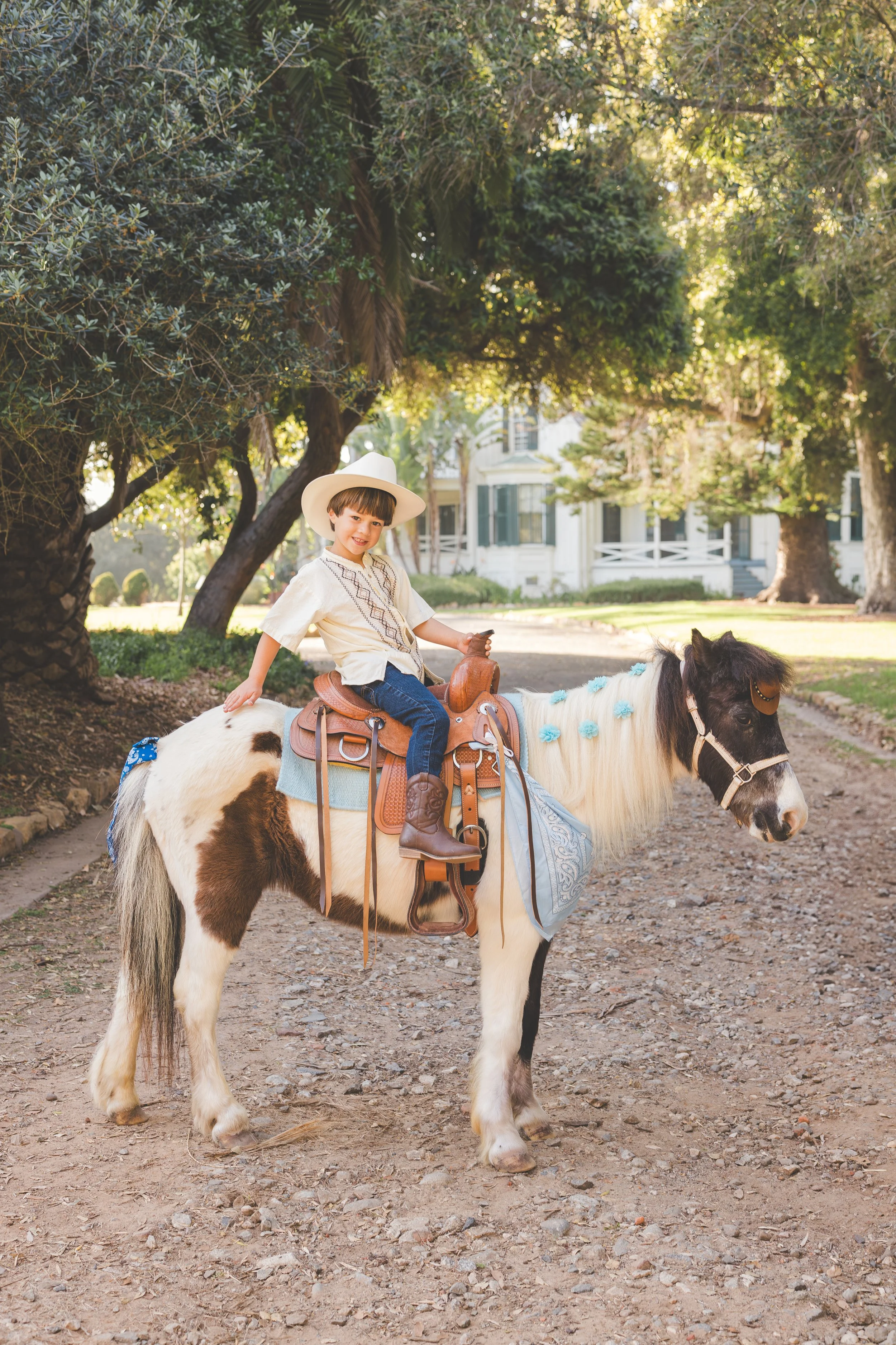 A young boy wearing a white cowboy hat and a cream-colored shirt riding a small pony with black and white fur. The pony has blue decorations on its mane and is standing on a dirt path with trees and a white house in the background.