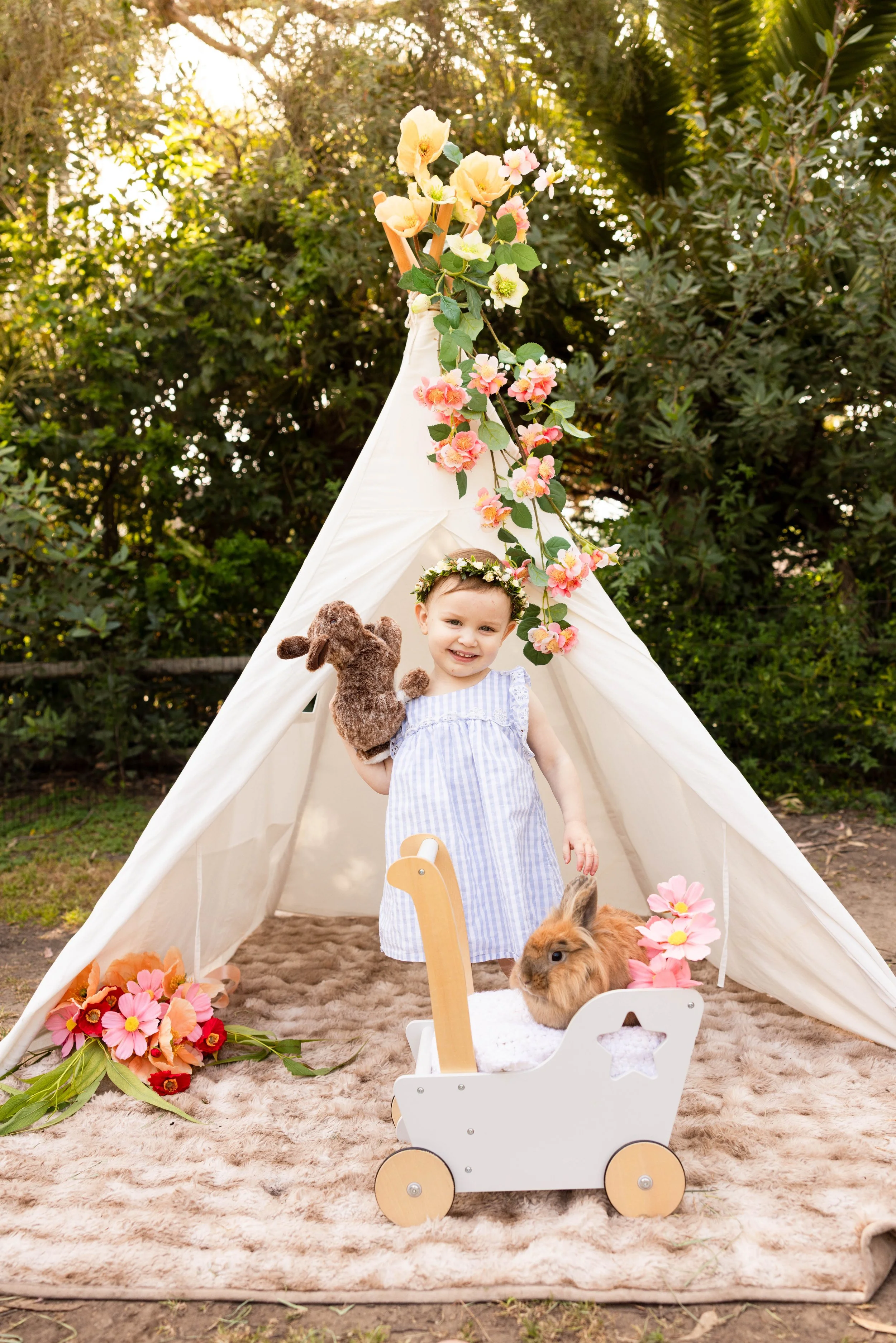 A young girl in a striped dress holding a stuffed animal inside a small decorative bed with a rabbit, surrounded by pink flowers, in front of a tent decorated with flowers, outdoors with trees in the background.