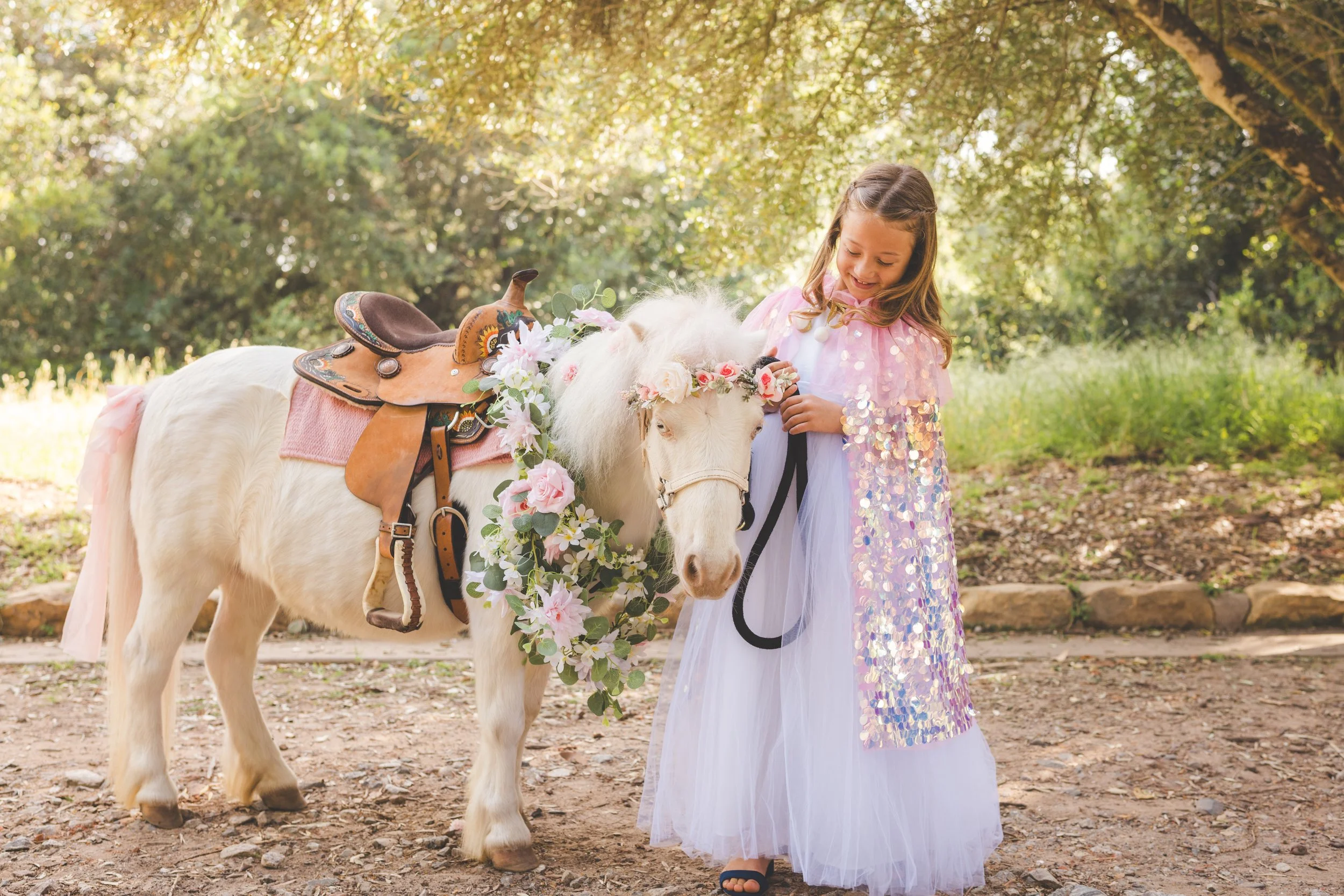 A young girl in a sparkly dress petting a white pony with a floral decoration on the saddle in a park with trees and sunlight.