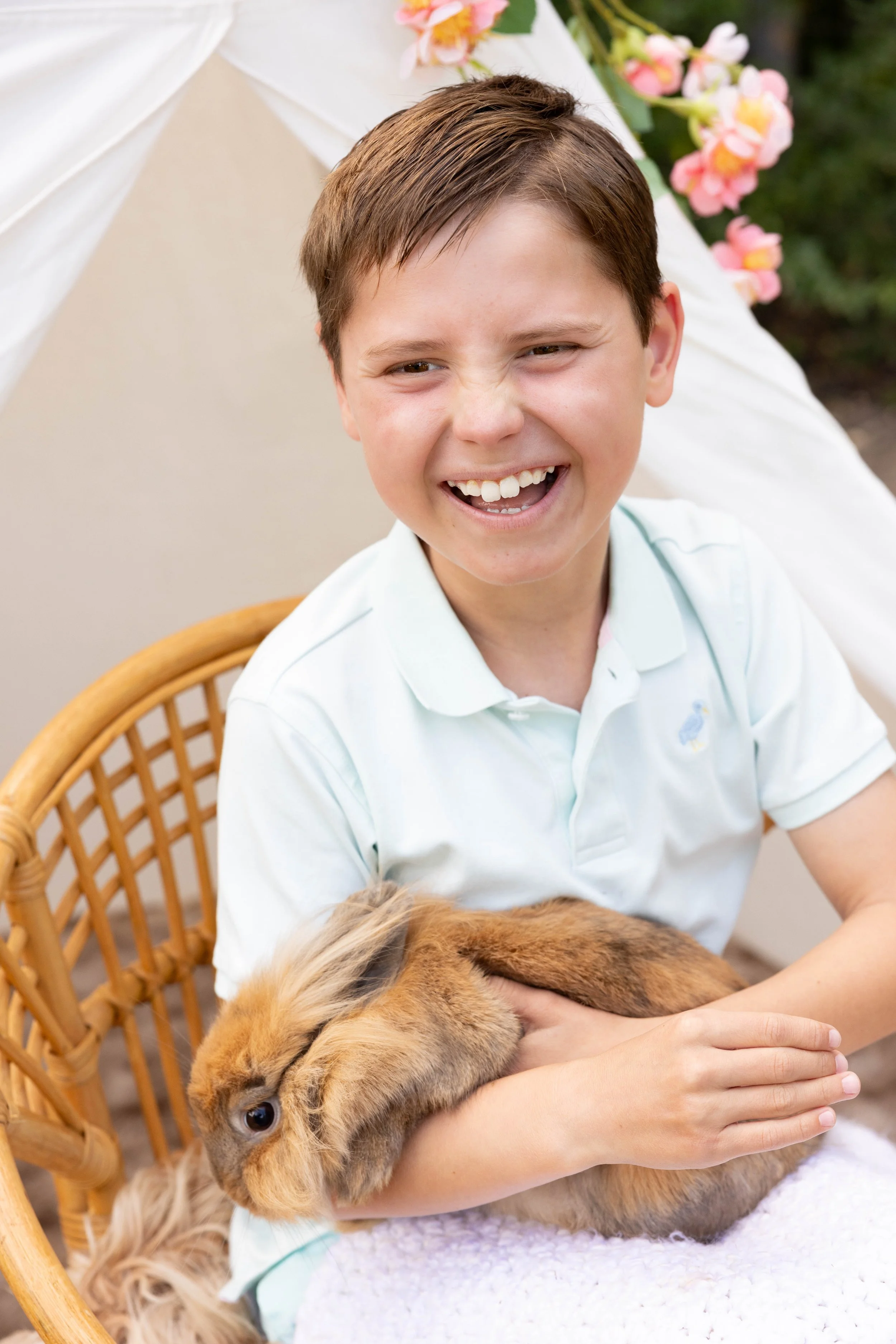 A young boy smiling happily while petting a brown rabbit on his lap, sitting on a wicker chair with pink flowers in the background.