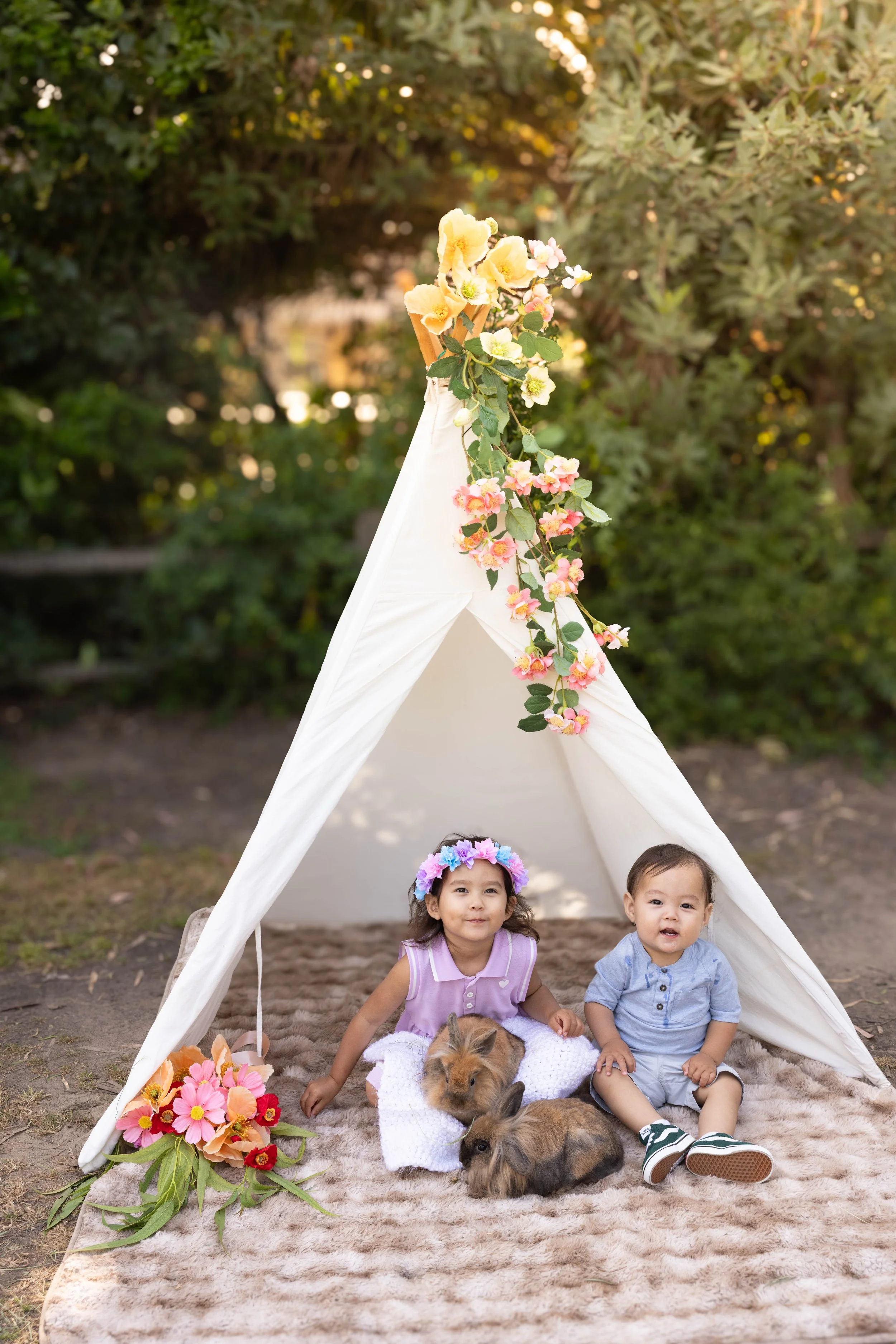Two young children, a girl and a boy, are sitting inside a small white tent decorated with pink, white, and purple flowers. The girl is wearing a purple dress and a flower crown, and the boy is in a light blue shirt and shorts. There are two rabbits,