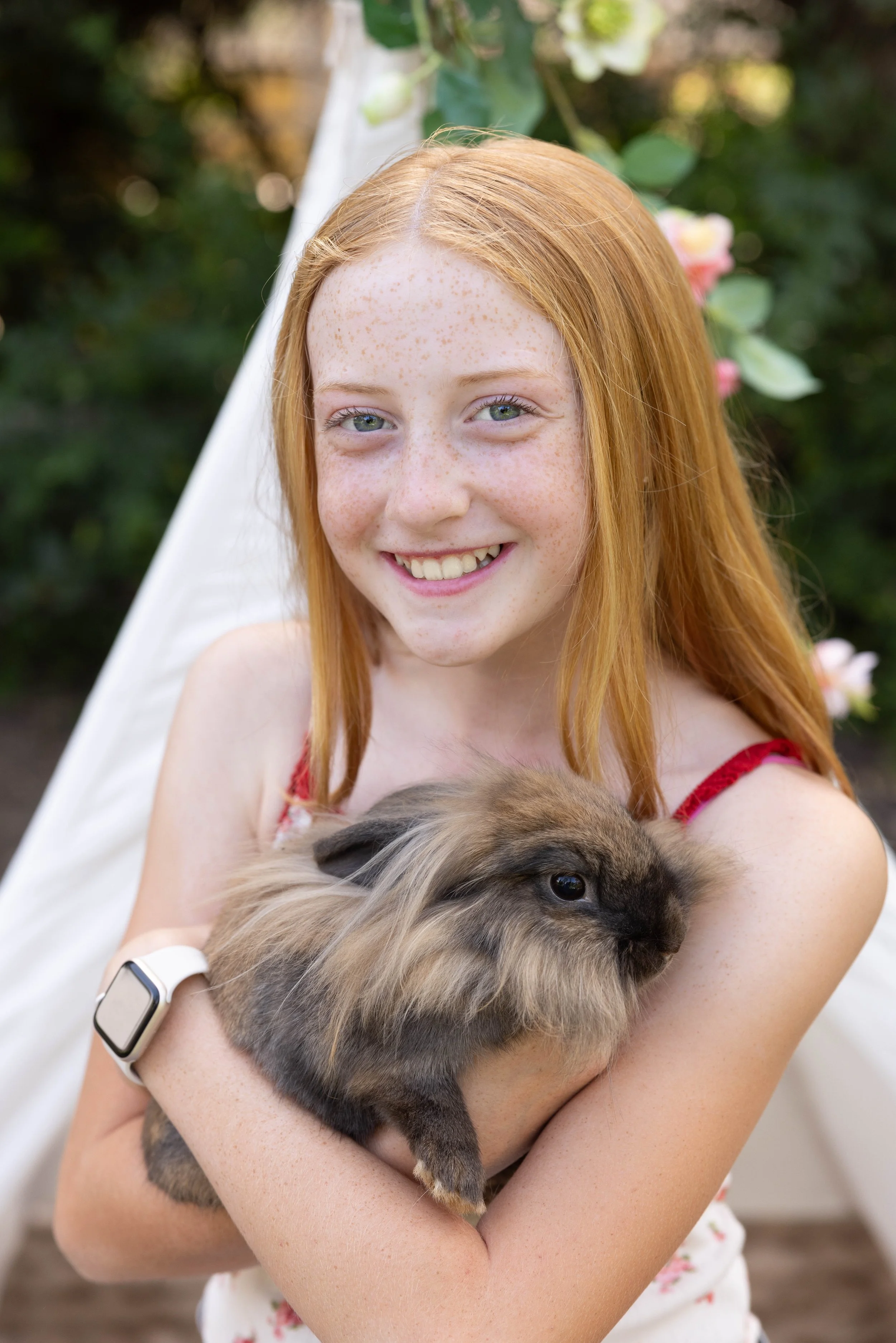 A young girl with red hair and freckles smiling while holding a fluffy rabbit outdoors. She wears a white wristband and a red dress with pink flower patterns.