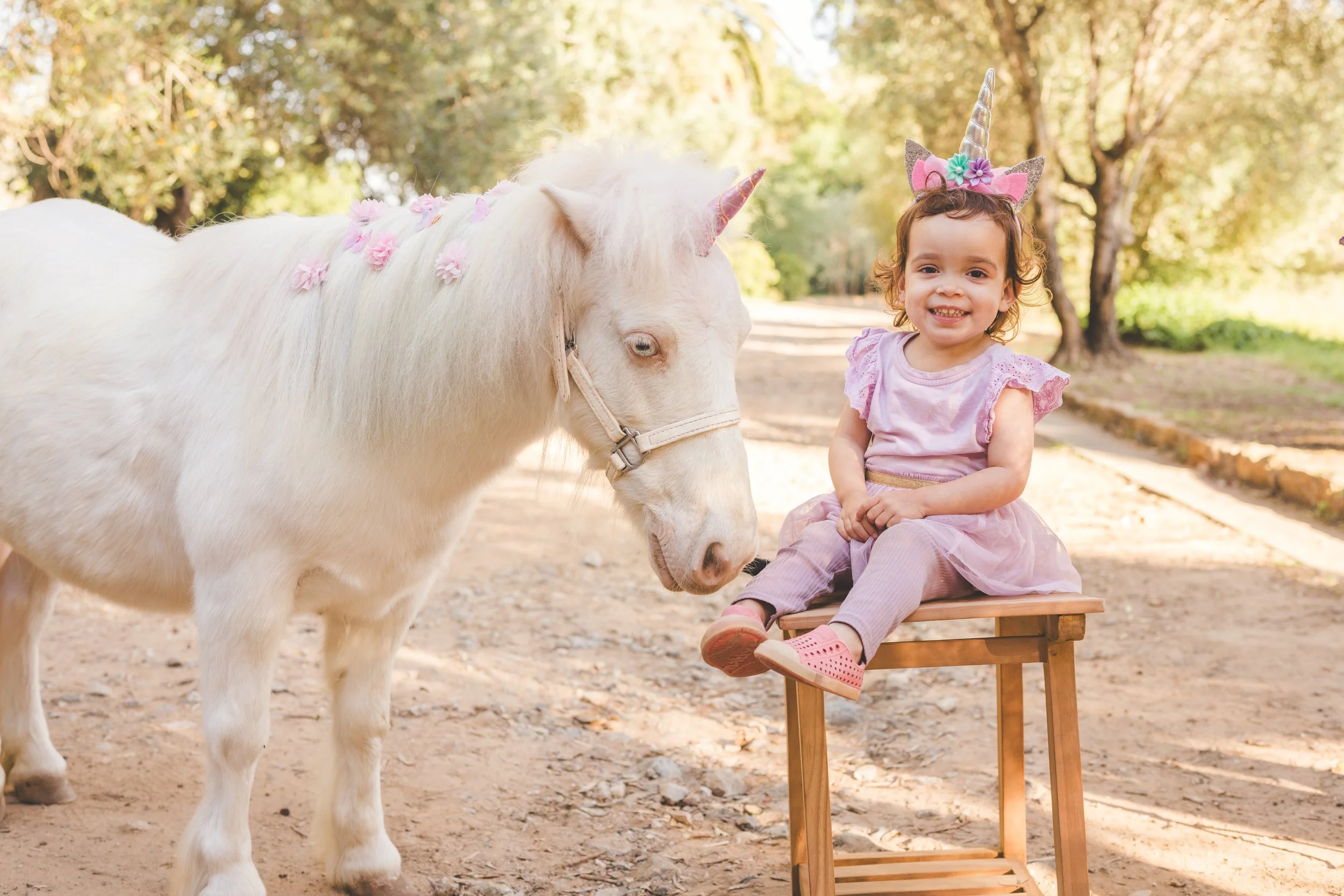 A young girl dressed as a unicorn, with a horn and colorful flowers on her head, sitting on a small wooden bench next to a white horse decorated with pink flowers on its mane, in a park with trees and sunlight in the background.