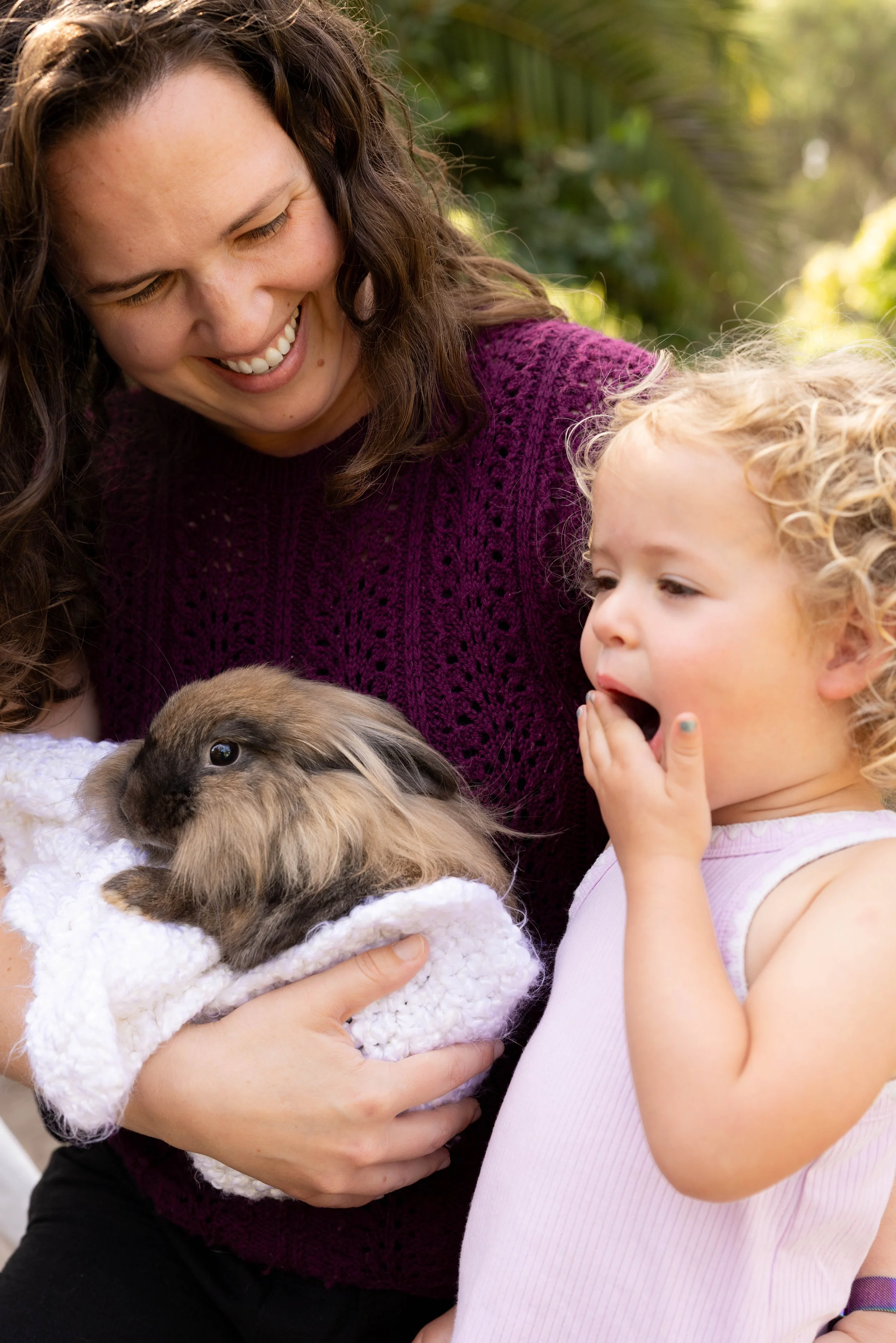 A woman with long brown hair smiling while holding a small, fluffy rabbit wrapped in a white blanket, with a young girl with curly blonde hair and a pink shirt observing the rabbit.